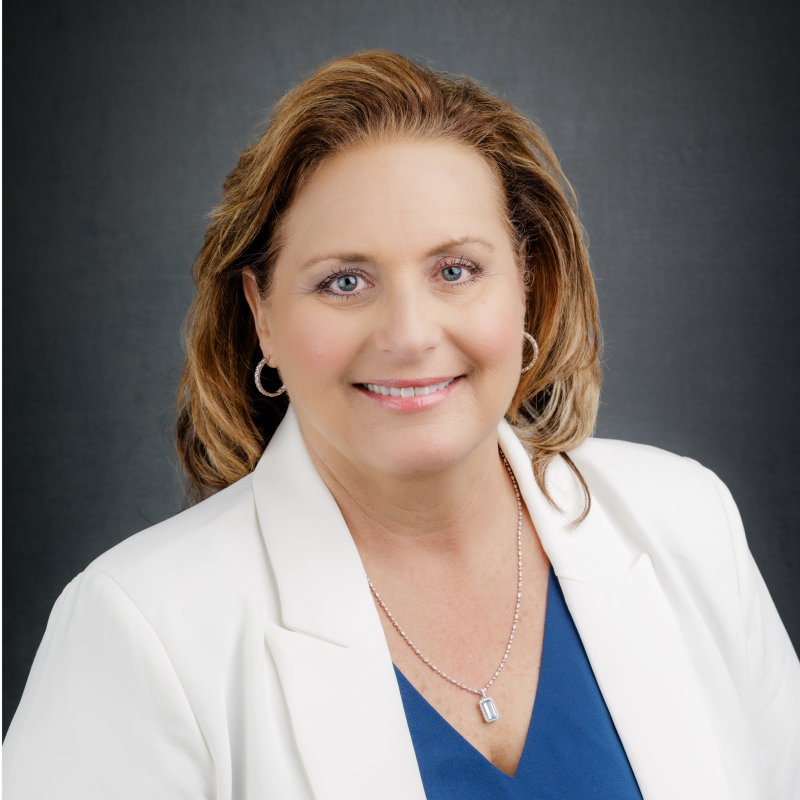 A middle-aged woman with light skin, shoulder-length auburn hair, and blue eyes, smiling and wearing a white blazer over a blue top, accessorized with hoop earrings and a necklace, against a dark background.