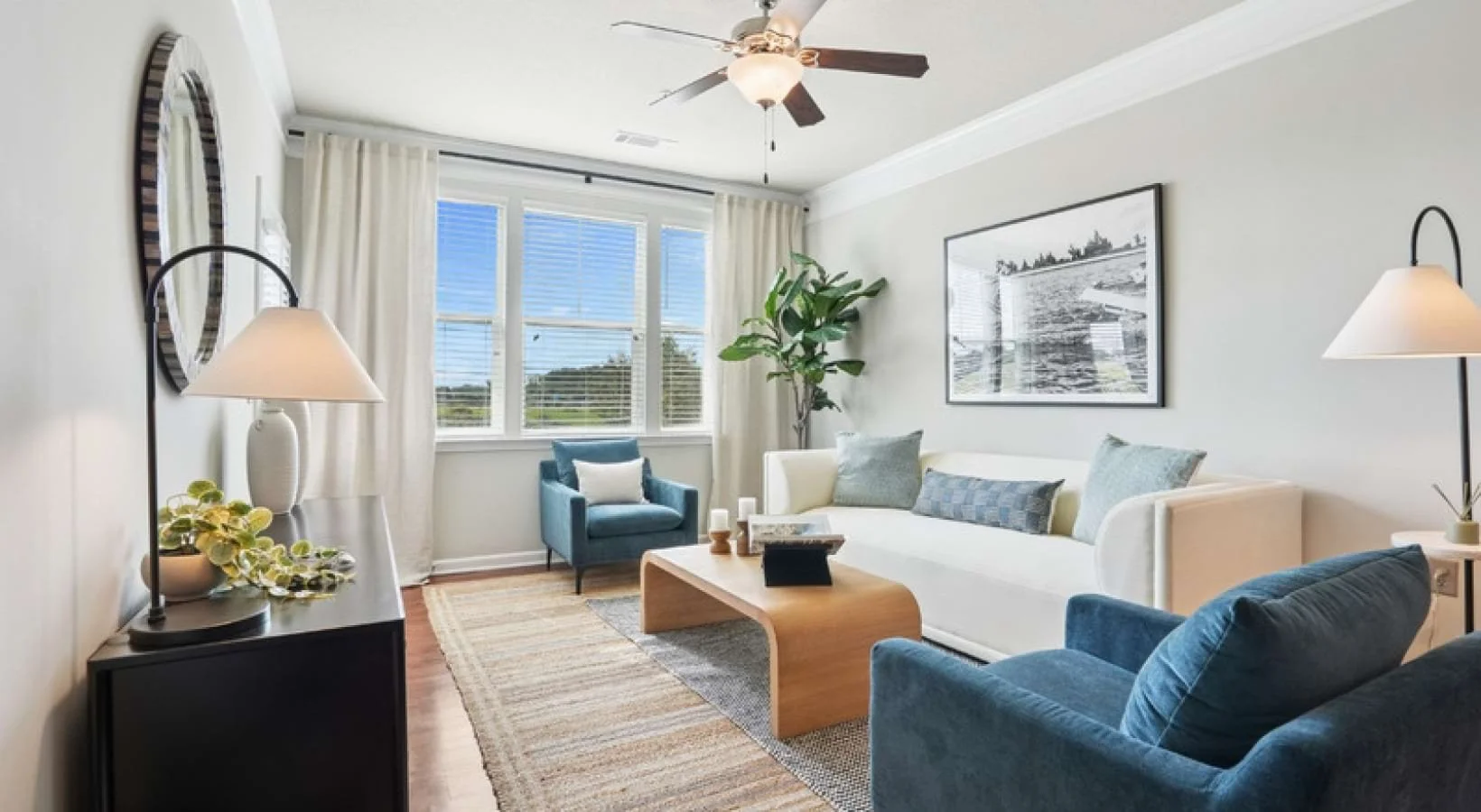 Bright living room with white walls, large window with blinds and curtains, white sofa with light blue and gray pillows, blue armchair, potted plant, framed black-and-white photograph, ceiling fan, and side table with lamp.