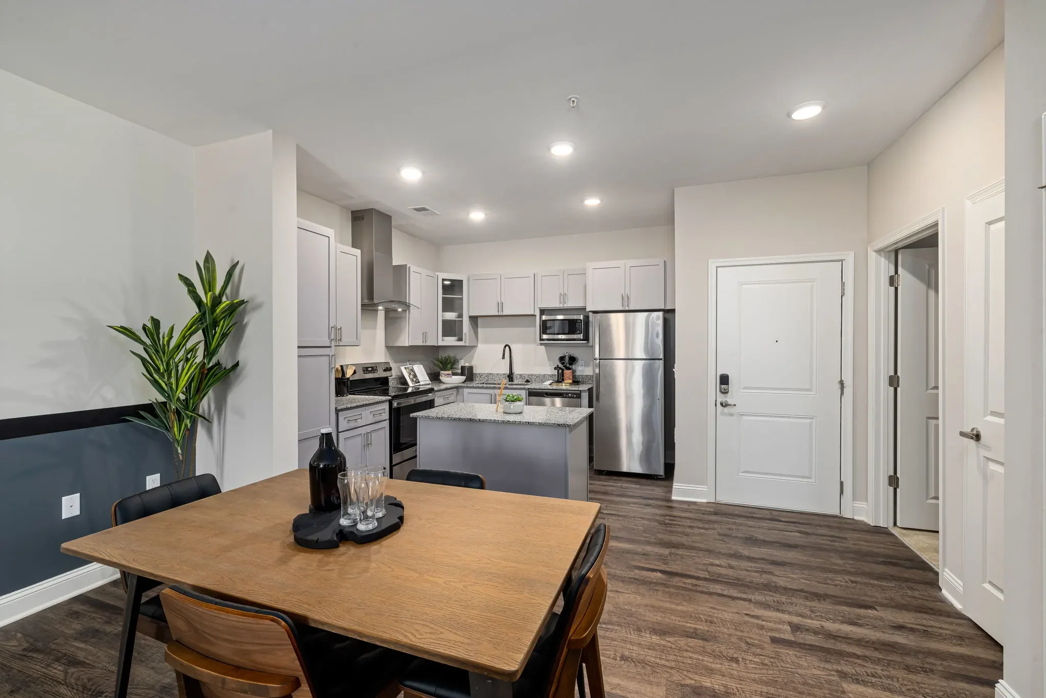 Modern kitchen with white cabinets, stainless steel refrigerator and microwave, granite countertops, black appliances, and a wooden dining table with chairs in the foreground.