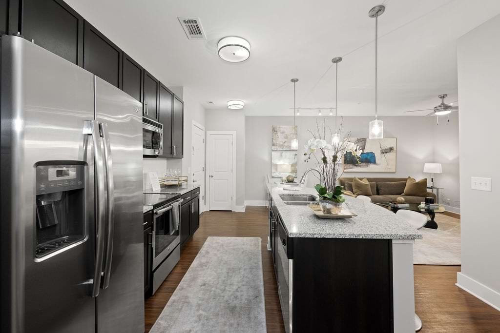 Modern kitchen with stainless steel refrigerator, microwave, and oven, dark upper cabinets, granite countertops, pendant lighting, and a living room with a sofa, coffee table, and artwork in the background.