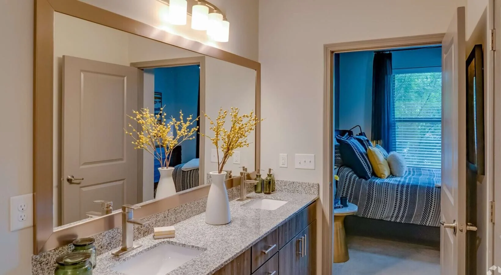 Bathroom vanity with mirror, flower vase, soap, toothbrush holder, and spa soap, leading to bedroom with window, blue curtains, and couch with yellow and patterned pillows.