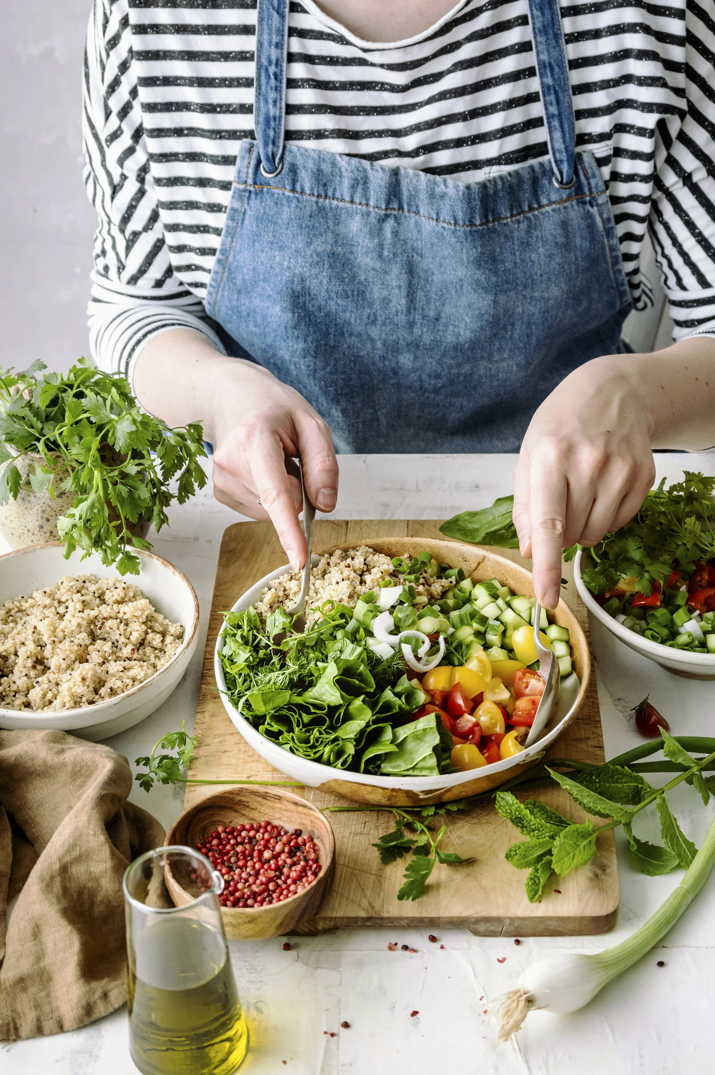 Lettuce, tomatoes, cucumber, spring onions, grains and herbs with nutritionist wearing striped top and denim apron putting two spoons in to toss