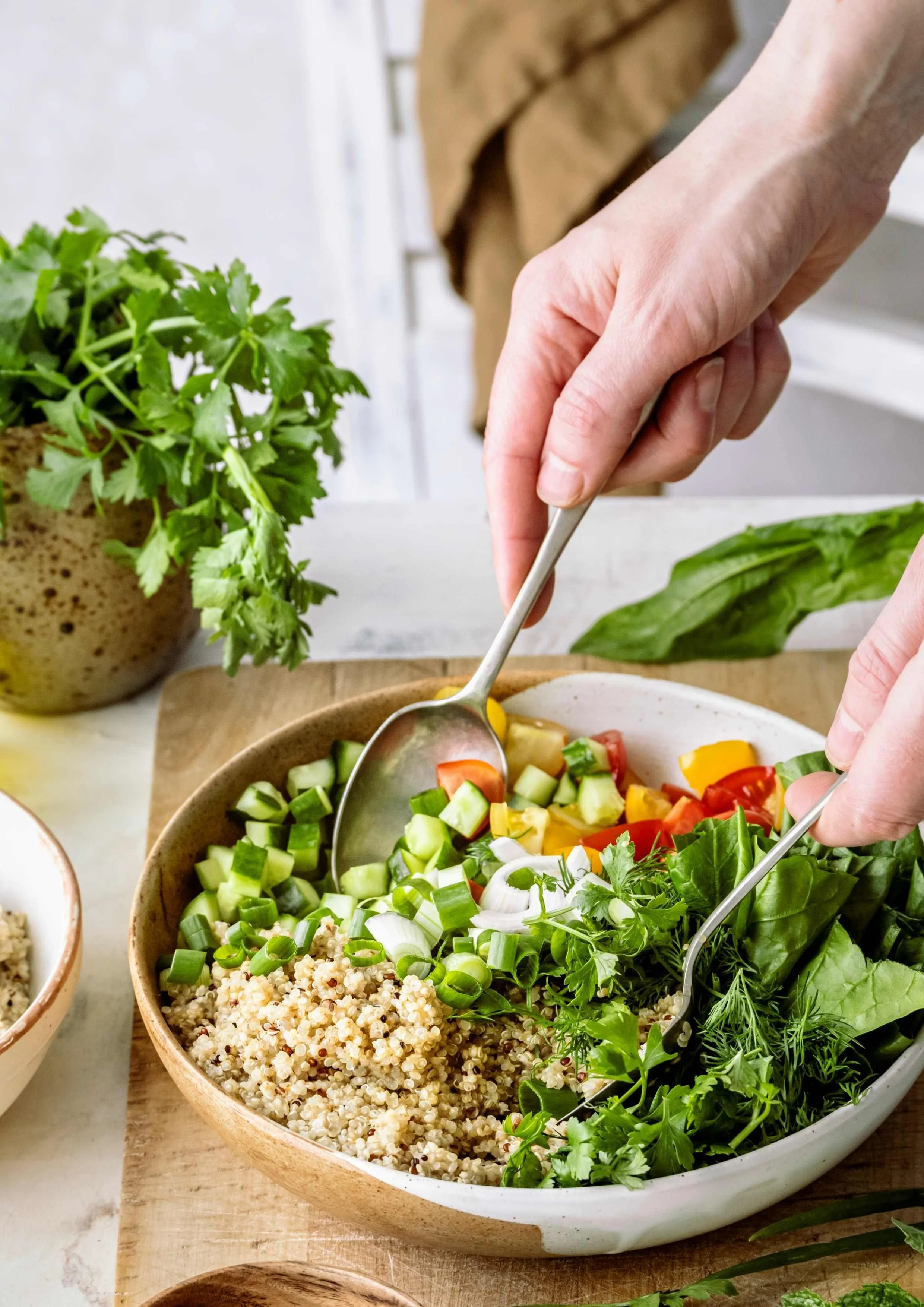 Salad in bowl with two hands using two spoons to toss grains, spring onions, lettuce, herbs, cucumber and tomatoes