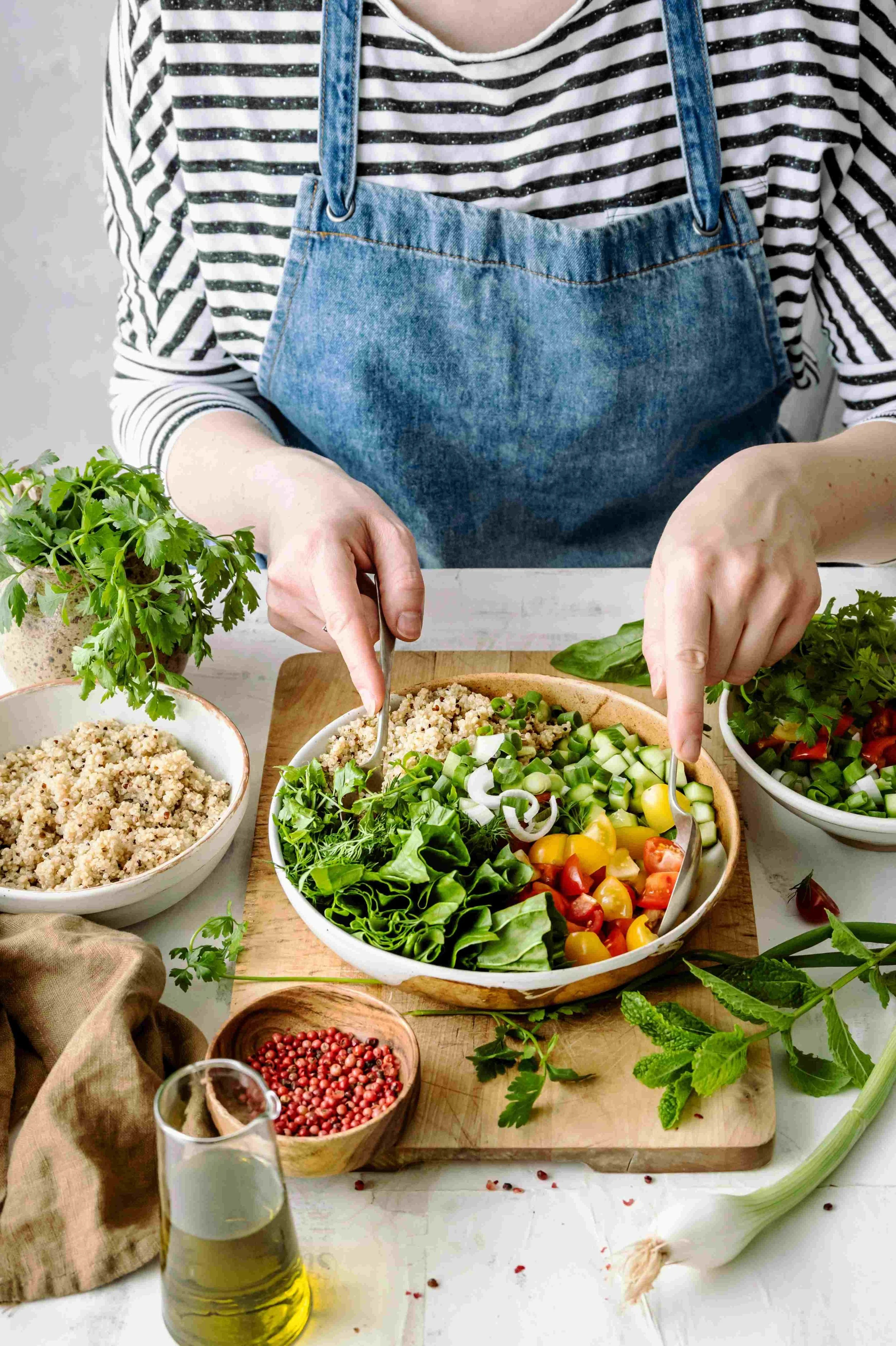 Lettuce, tomatoes, cucumber, spring onions, grains and herbs with person wearing striped top and denim apron putting two spoons in to toss