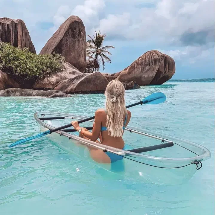 A woman with long blonde hair in a blue bikini sitting in a kayak on clear turquoise water near rocky islands with palm trees.