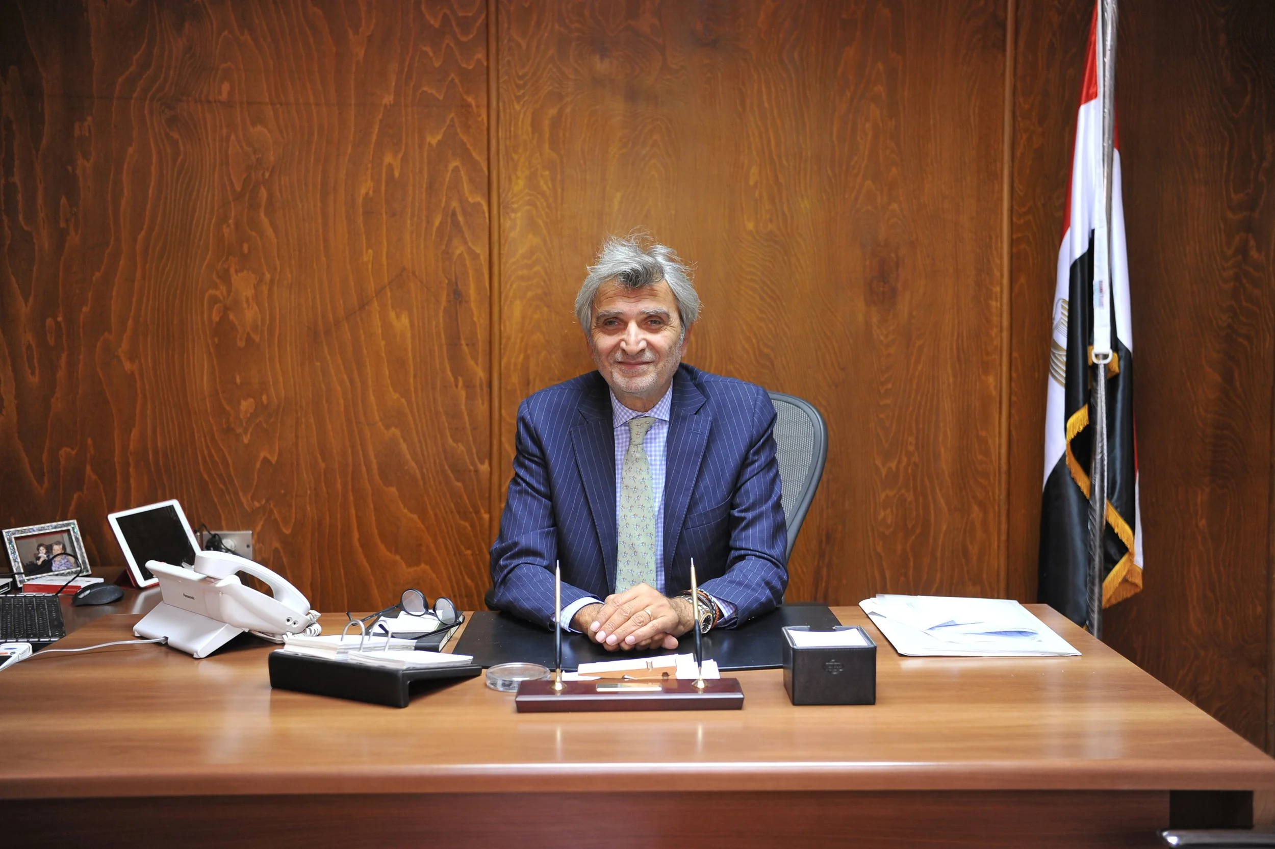 A middle-aged man with gray hair and a mustache, dressed in a navy pinstripe suit and patterned tie, sitting at a large wooden desk in an office with wooden-paneled walls. The desk has various electronic devices, glasses, a calling device, and papers. To the right, there are framed photos and a landline phone. Behind him, on the right side, there are flags, likely representing the country and organization.