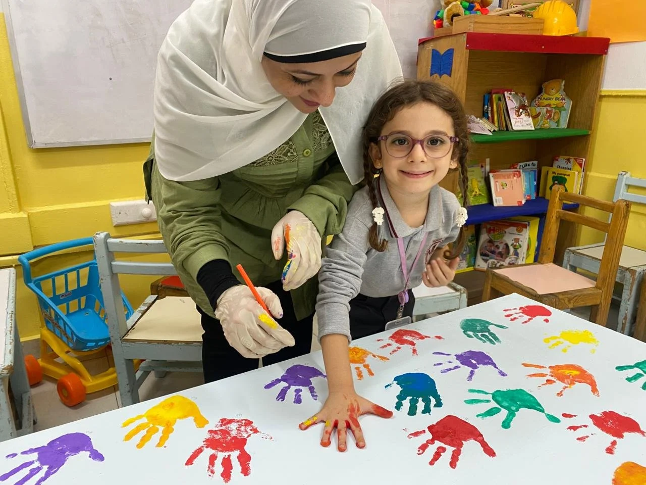 A young girl with glasses smiling as she shows her painted handprint on a table covered with colorful handprint art. An adult woman standing next to her is painting another handprint. The background features children’s books and toys.