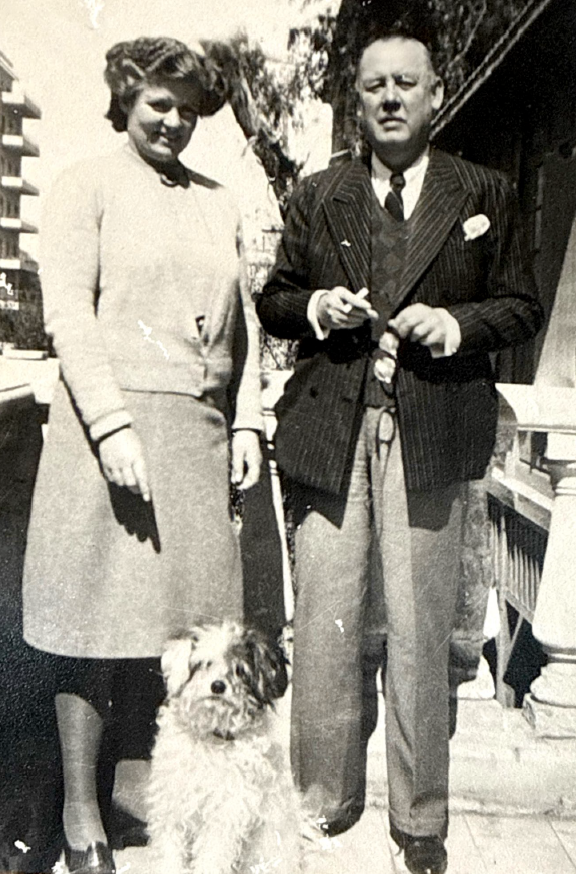 Black and white photo of a woman and a man standing outdoors with a dog at their feet. The woman is wearing a skirt and sweater, and the man is in a suit and tie holding something in his hand. Trees and buildings are visible in the background.