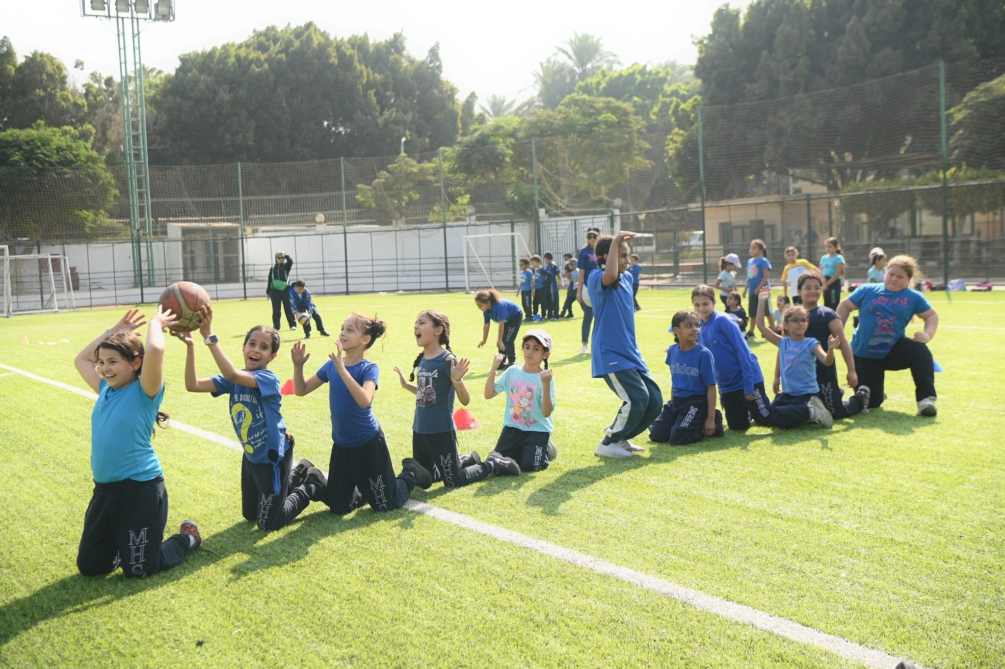 Children in blue sports uniforms participating in a field activity on a sunny day at a sports field, with some kneeling and others standing, among trees and a sports fence.
