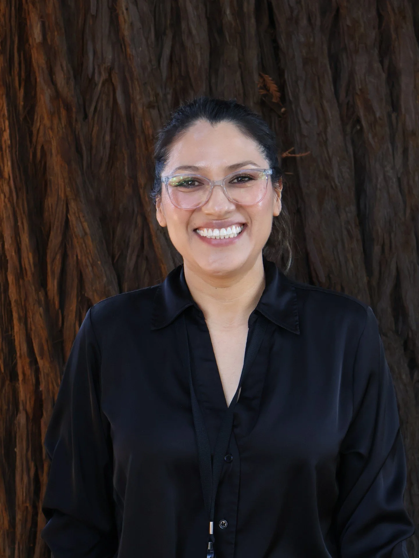 A woman with dark hair pulled back, wearing clear glasses and a black top, standing in front of a large tree trunk, smiling broadly.