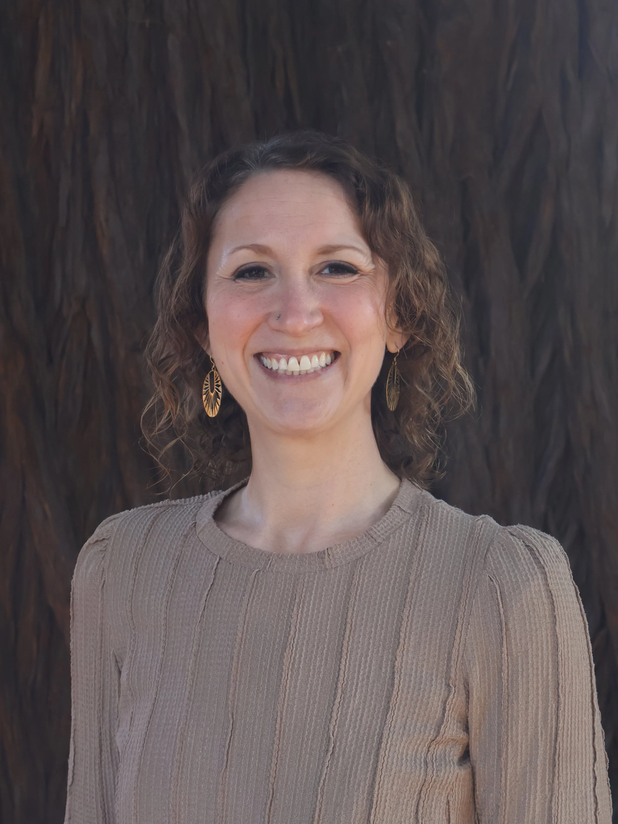 A woman with curly brown hair and earrings smiling in front of a dark wooden background.