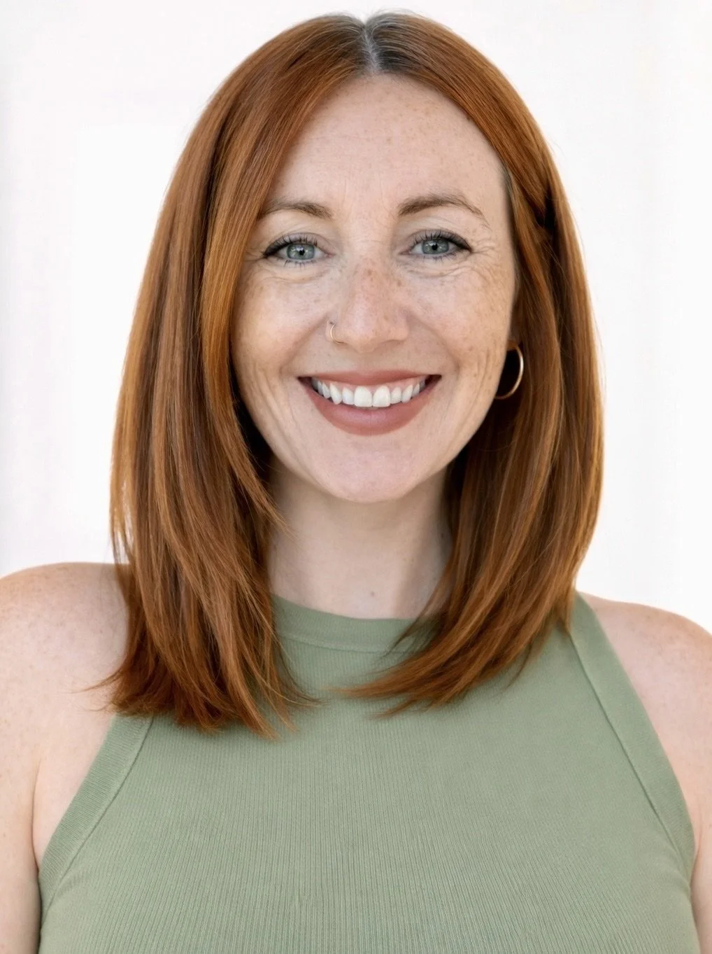 A smiling red-haired woman with blue eyes wearing a sleeveless green top and hoop earrings, facing the camera against a white background.