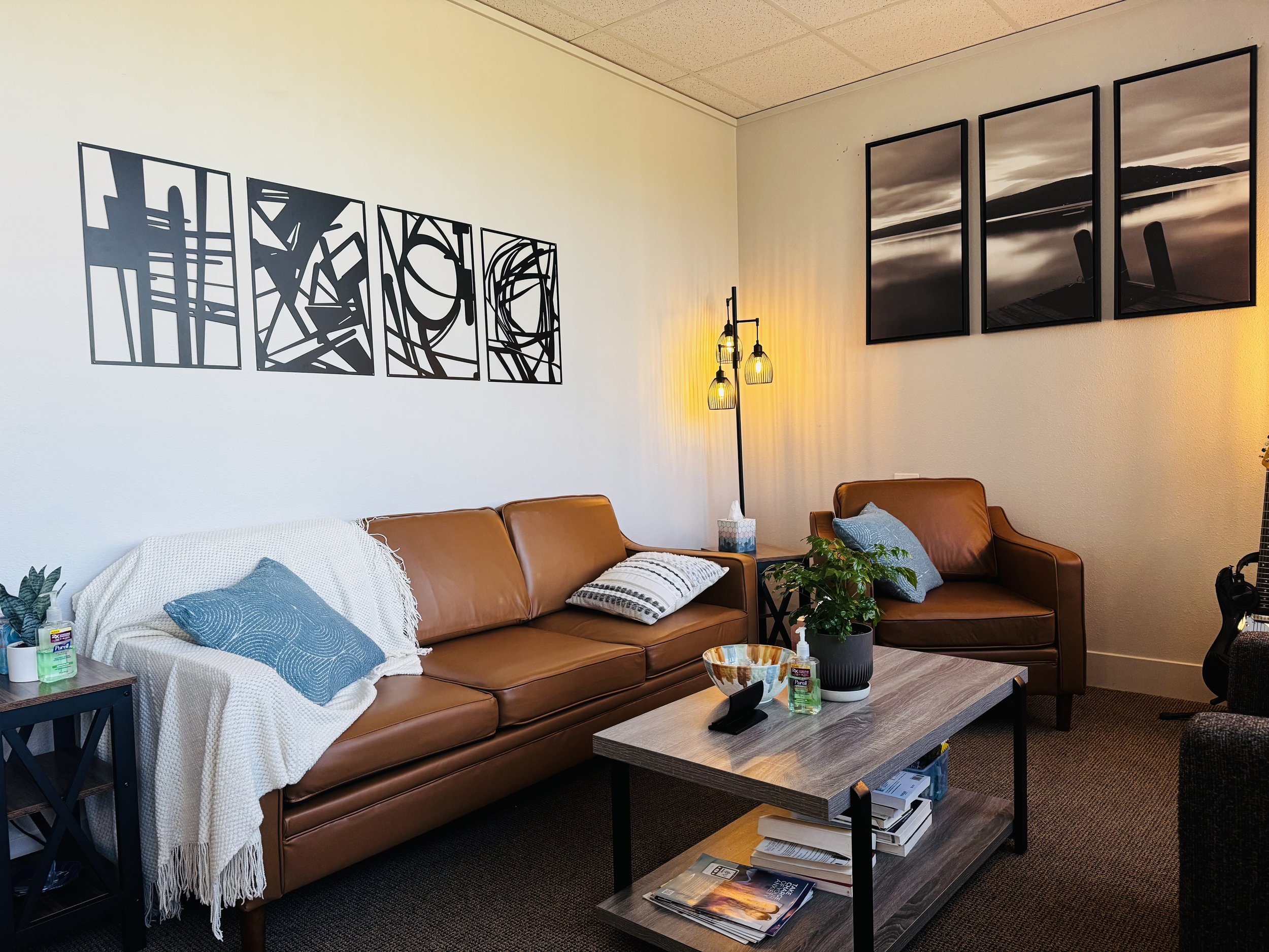 Living room with a leather sofa, decorative pillows, a throw blanket, artwork on the walls, a coffee table with magazines, a lamp with hanging lights, and various items including hand sanitizer and tissues.