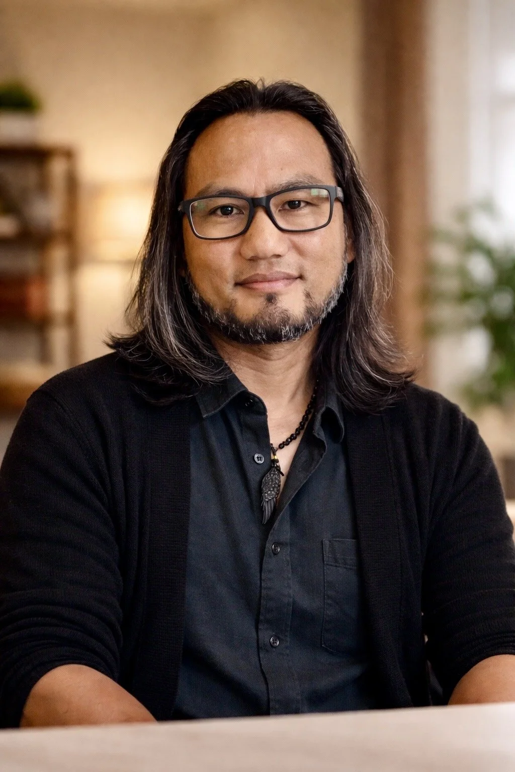 A man with long dark hair, a beard, and glasses sitting indoors with a blurred background of a room with warm lighting, shelves, and plants.