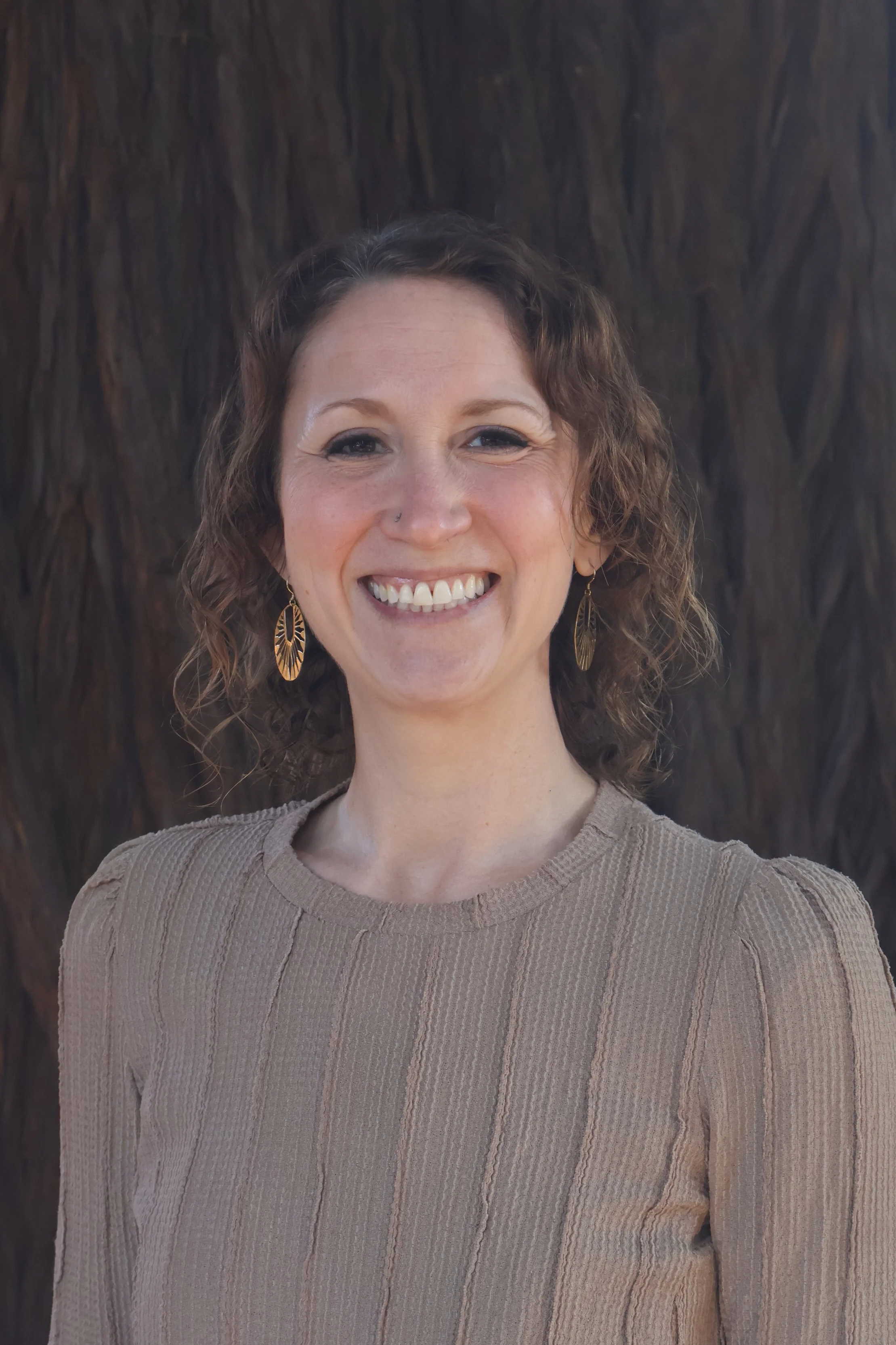 A woman with curly brown hair, wearing gold earrings and a beige ribbed sweater, smiling in front of a dark textured background.