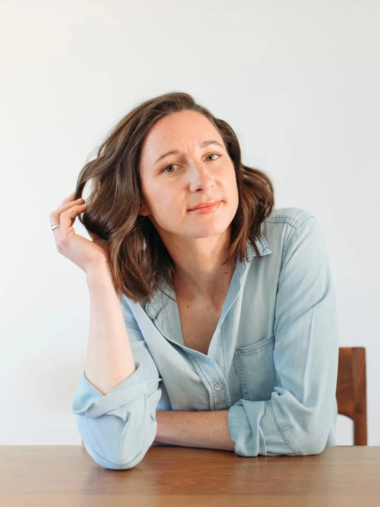A woman with shoulder-length brown hair wearing a light blue shirt sitting at a wooden table with a plain white background.