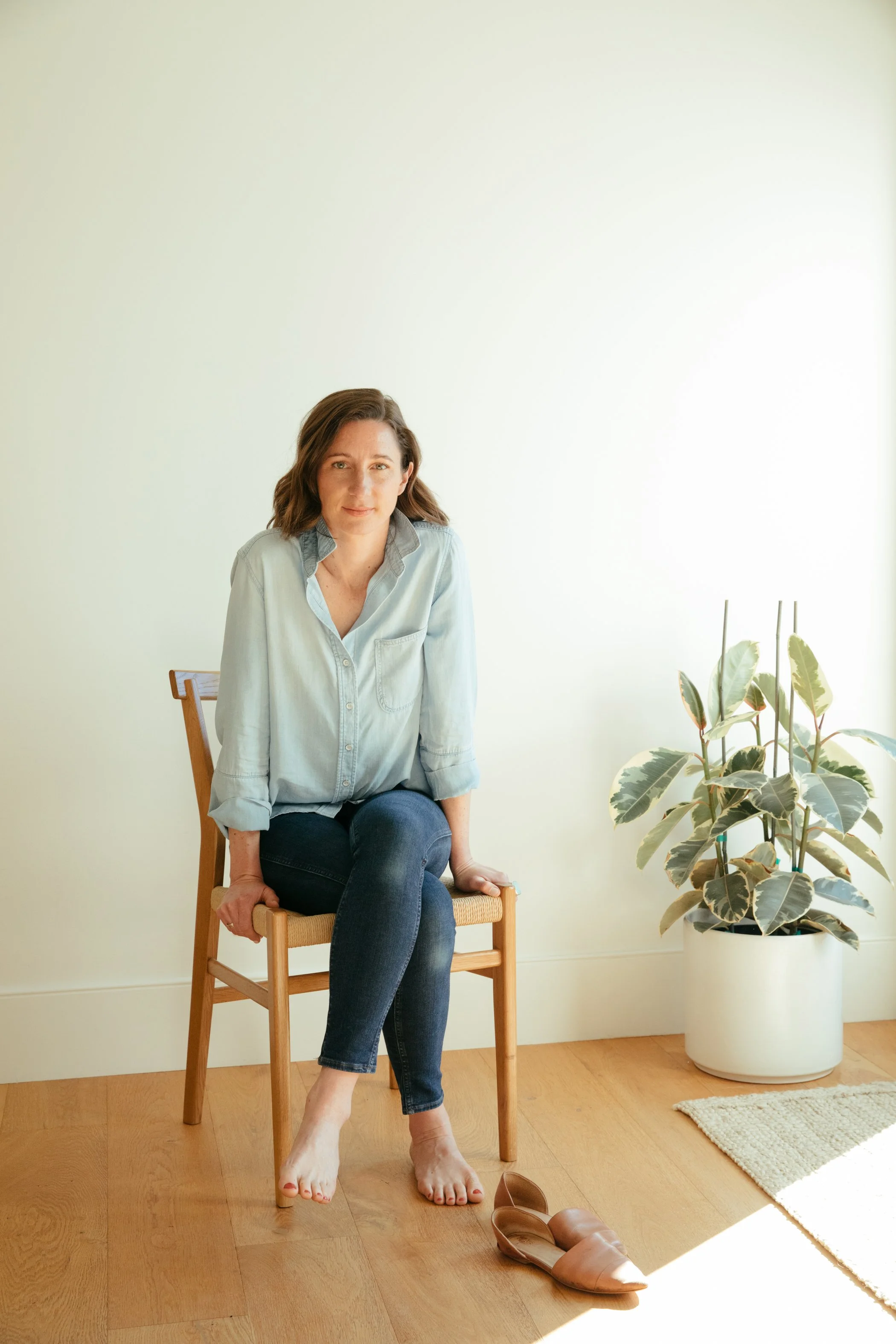 A woman with shoulder-length brown hair sitting on a wooden chair in a room with a potted plant beside her. She is wearing a light denim shirt and dark jeans, with her feet touching the wooden floor. There are brown slip-on shoes and a pair of glasses on the floor nearby. The room has a light-colored wall, a beige rug, and a window casting natural light.