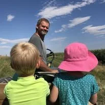 A man with two children outdoors on a sunny day, one child wearing a pink hat and the other in a yellow shirt, with blue sky and clouds in the background.