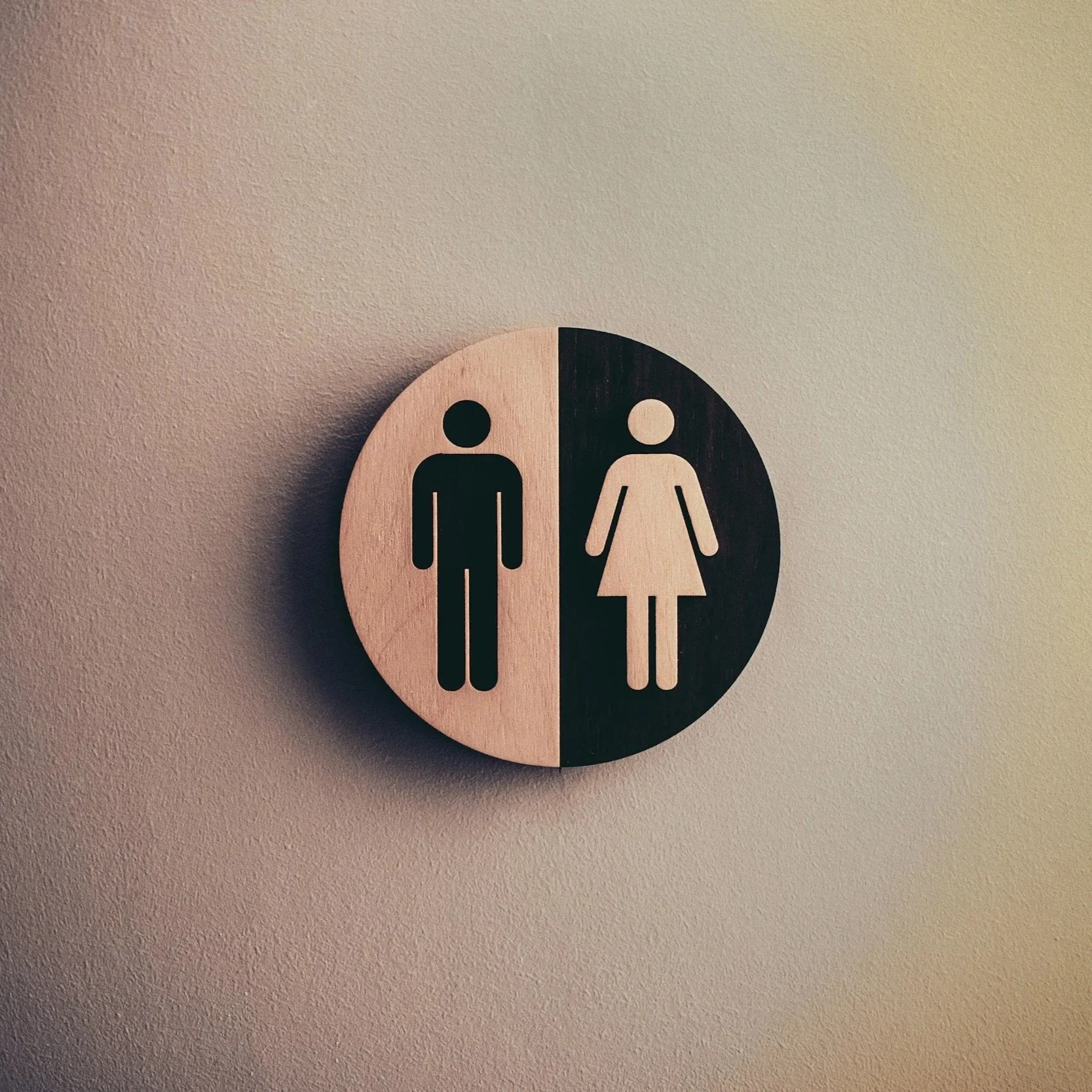 A circular, two-tone sign with male and female restroom symbols, half in light wood color and half in dark wood color, mounted on a beige wall.