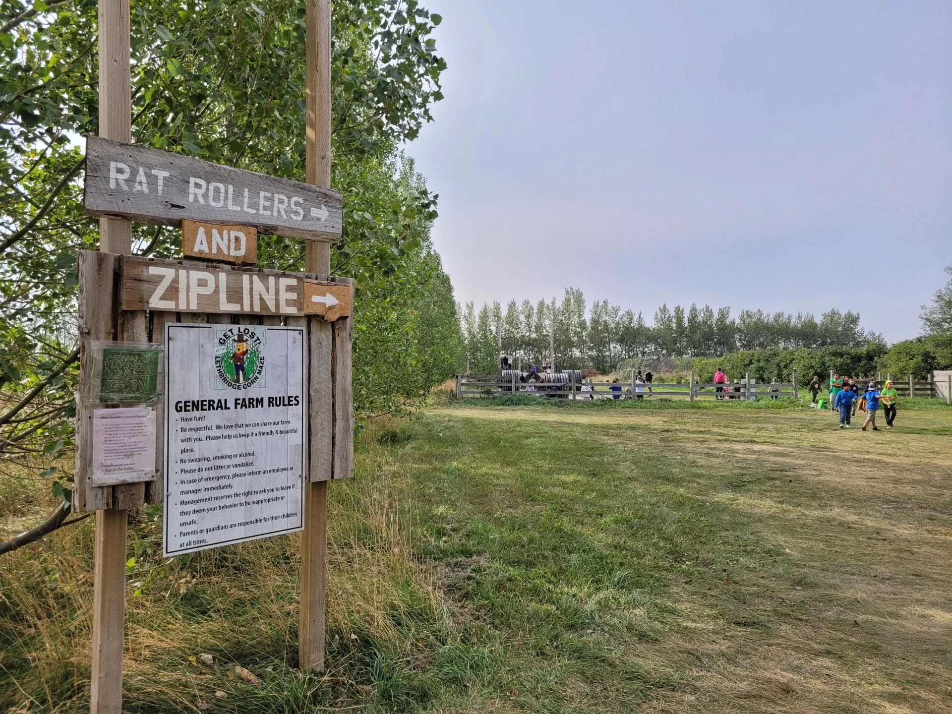 A sign at a farm entrance indicates directions to rat rollers and zipline, with a background of people walking and activities in a rural setting, surrounded by trees and a cloudy sky.