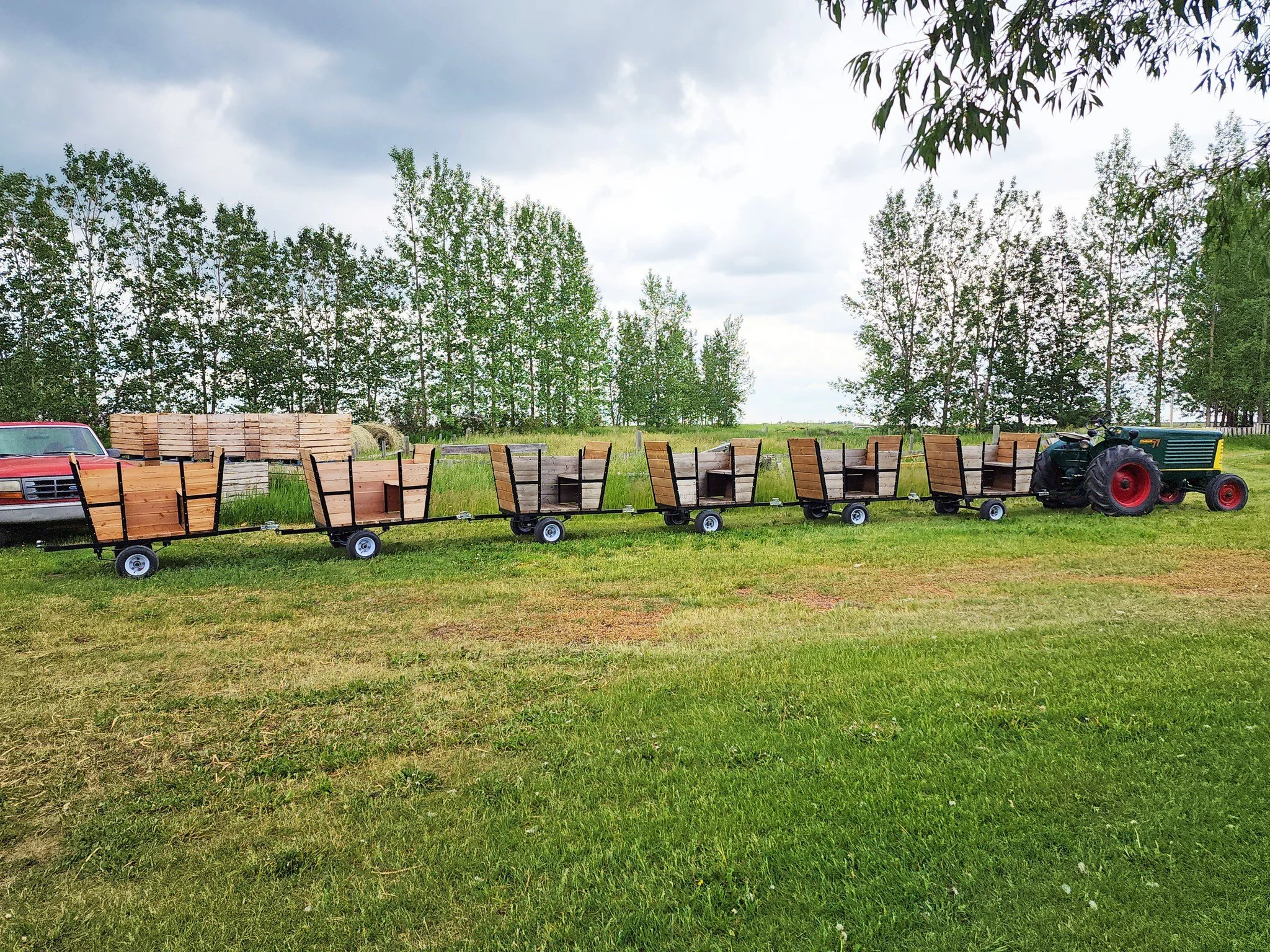 A tractor hauling wooden carts on a grassy field with trees and cloudy sky in the background.