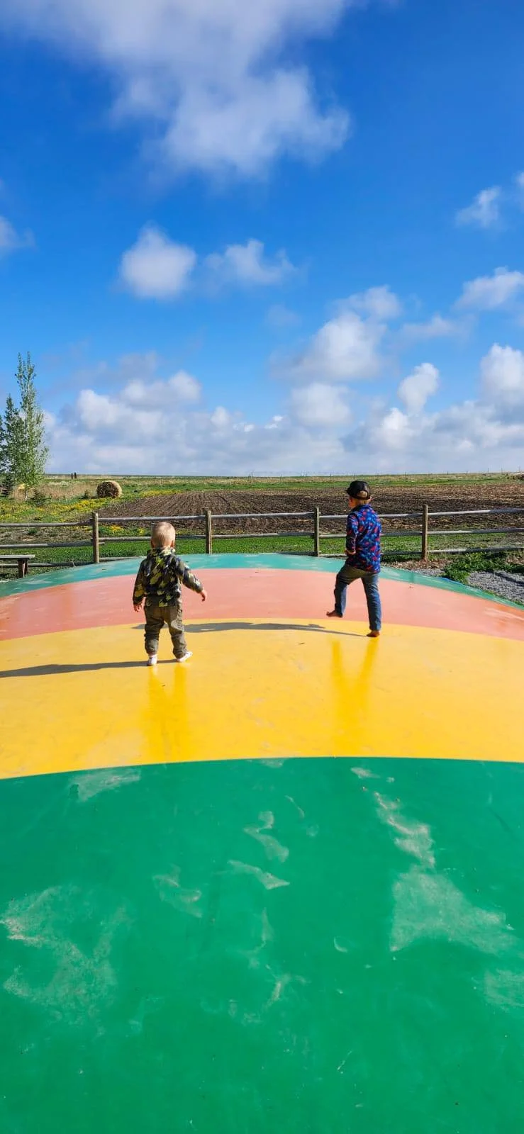 Two children playing on a jumping pillow under a blue sky with scattered clouds, with fields and a fence in the background.