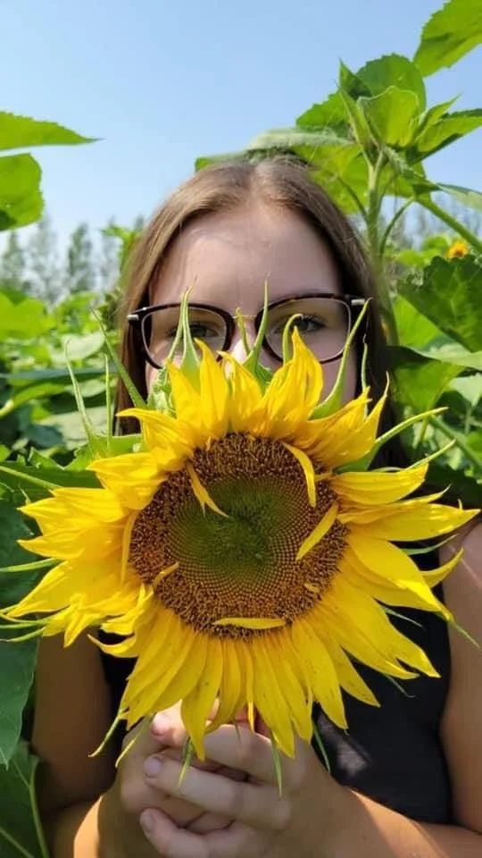 A woman with glasses holding a large sunflower in front of her face in a sunflower field.