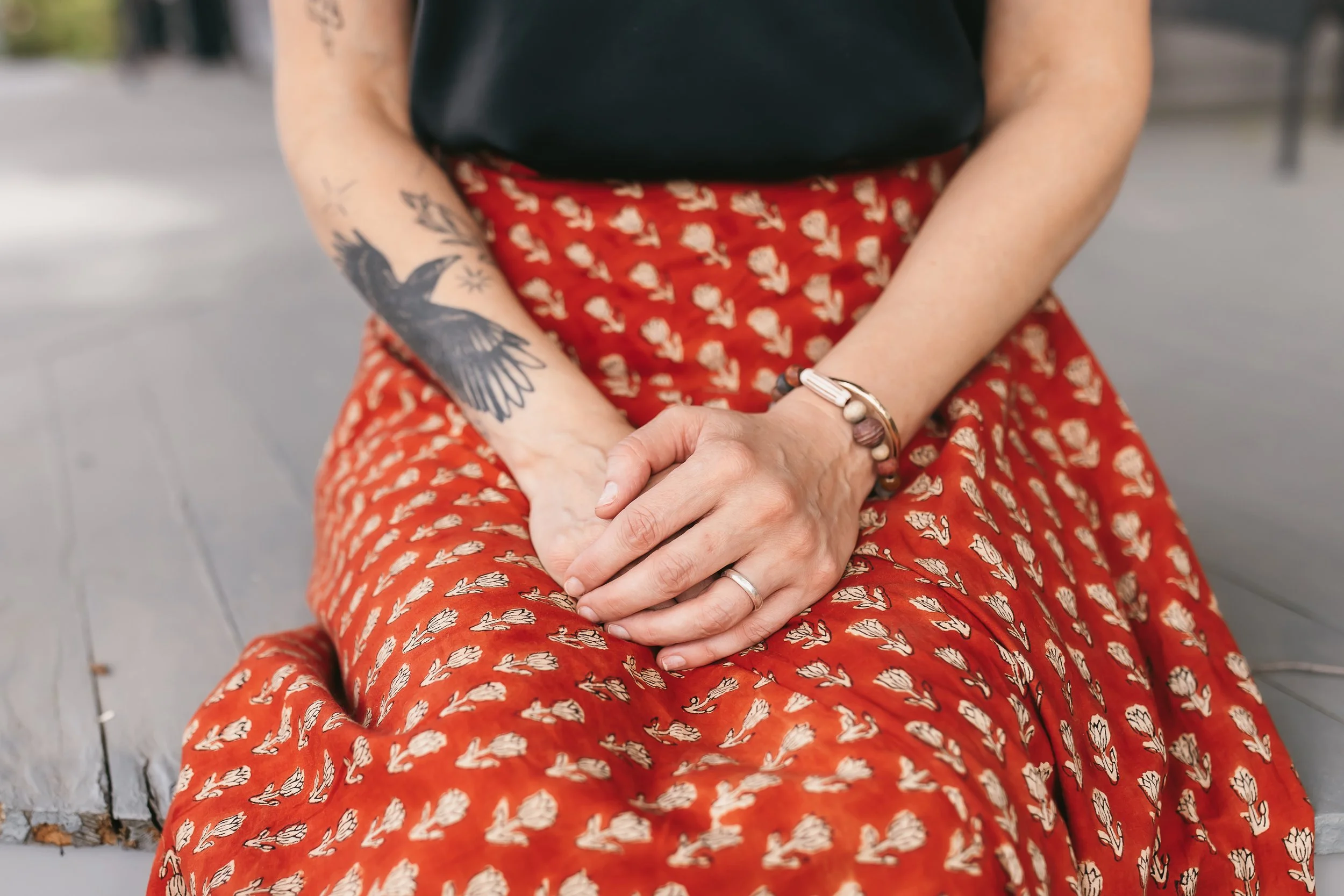 Close-up of a woman sitting cross-legged on a bench, wearing a black shirt and red patterned skirt, with tattoos on her arm and various bracelets on her wrist.