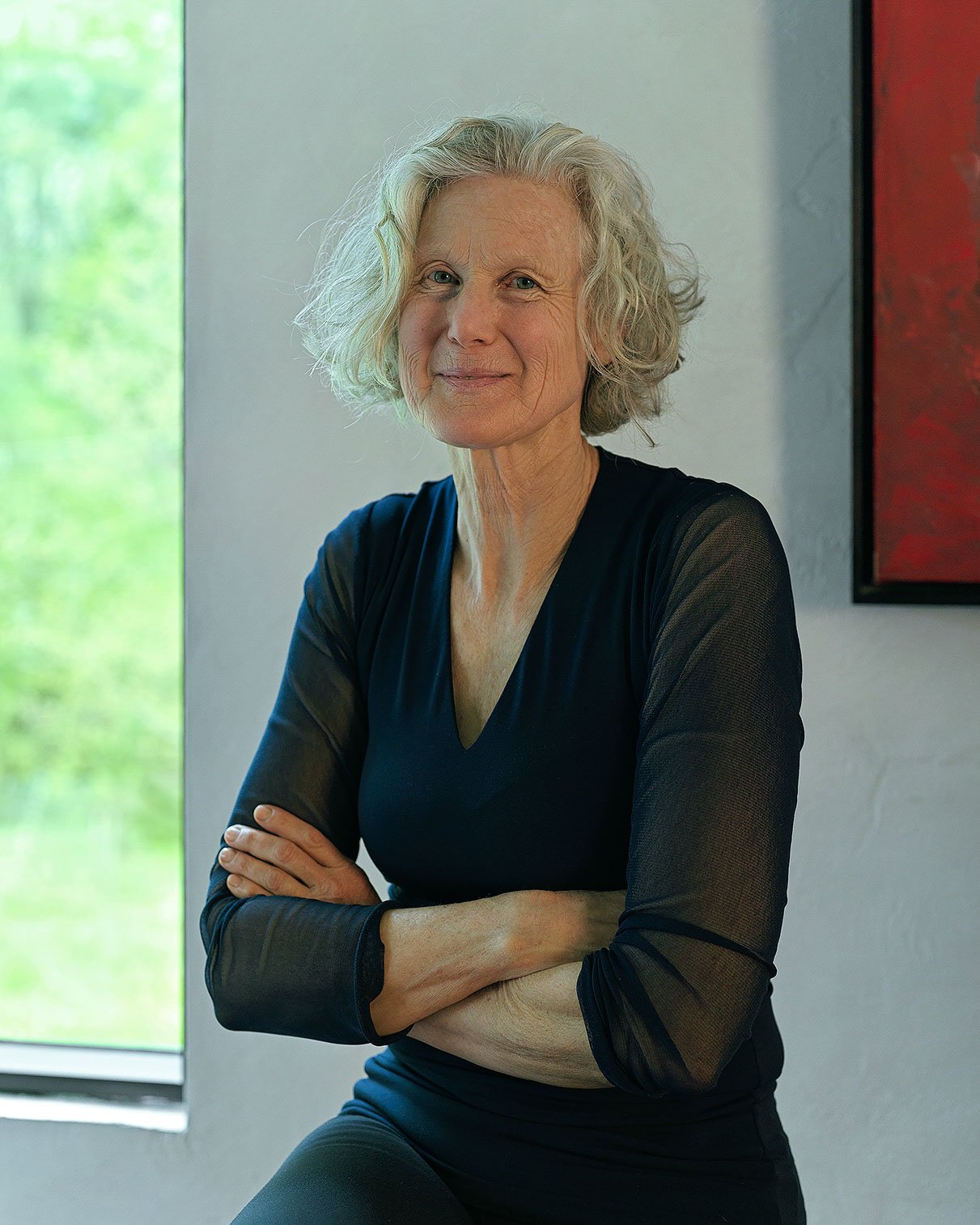 Older woman with curly gray hair, wearing a black sheer long-sleeve top, standing with arms crossed, smiling slightly, indoors near a window with green scenery outside.