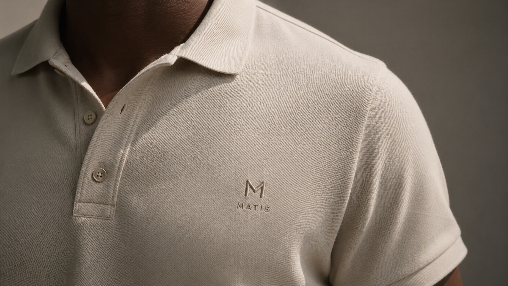 Nahaufnahme eines Mannes in einem beige Polohemd mit Logo "Matis" auf der Brust, schwarzer Haut und dunkler Haut. Der Hintergrund ist grau.