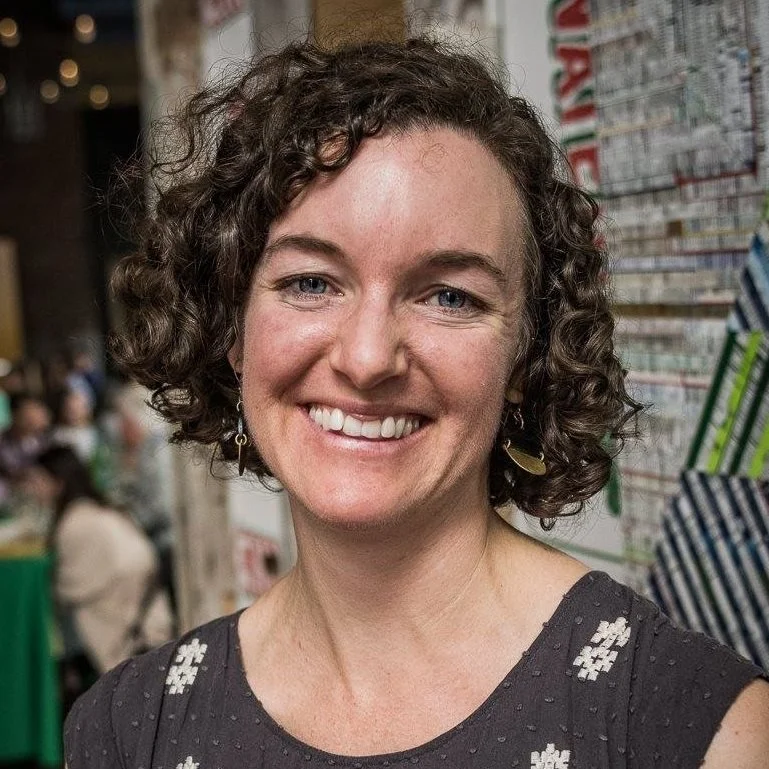 A woman with curly brown hair smiling, wearing earrings and a black shirt with a floral pattern, standing in an indoor setting with blurred background of people and colorful items on a wall.