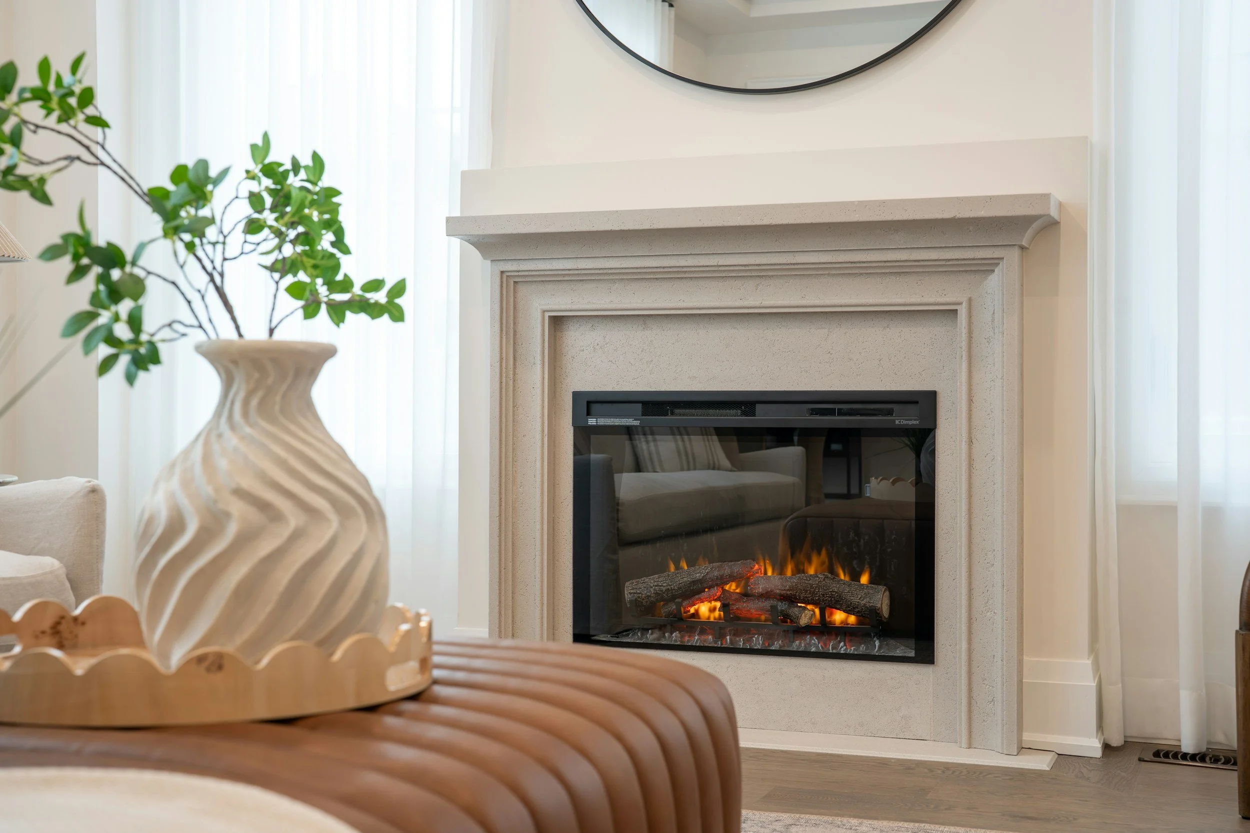 A living room with a beige fireplace, a round mirror above it, and light-colored curtains. There is a large beige vase with a green leafy plant. Part of a patterned wooden table or tray is visible in the foreground.