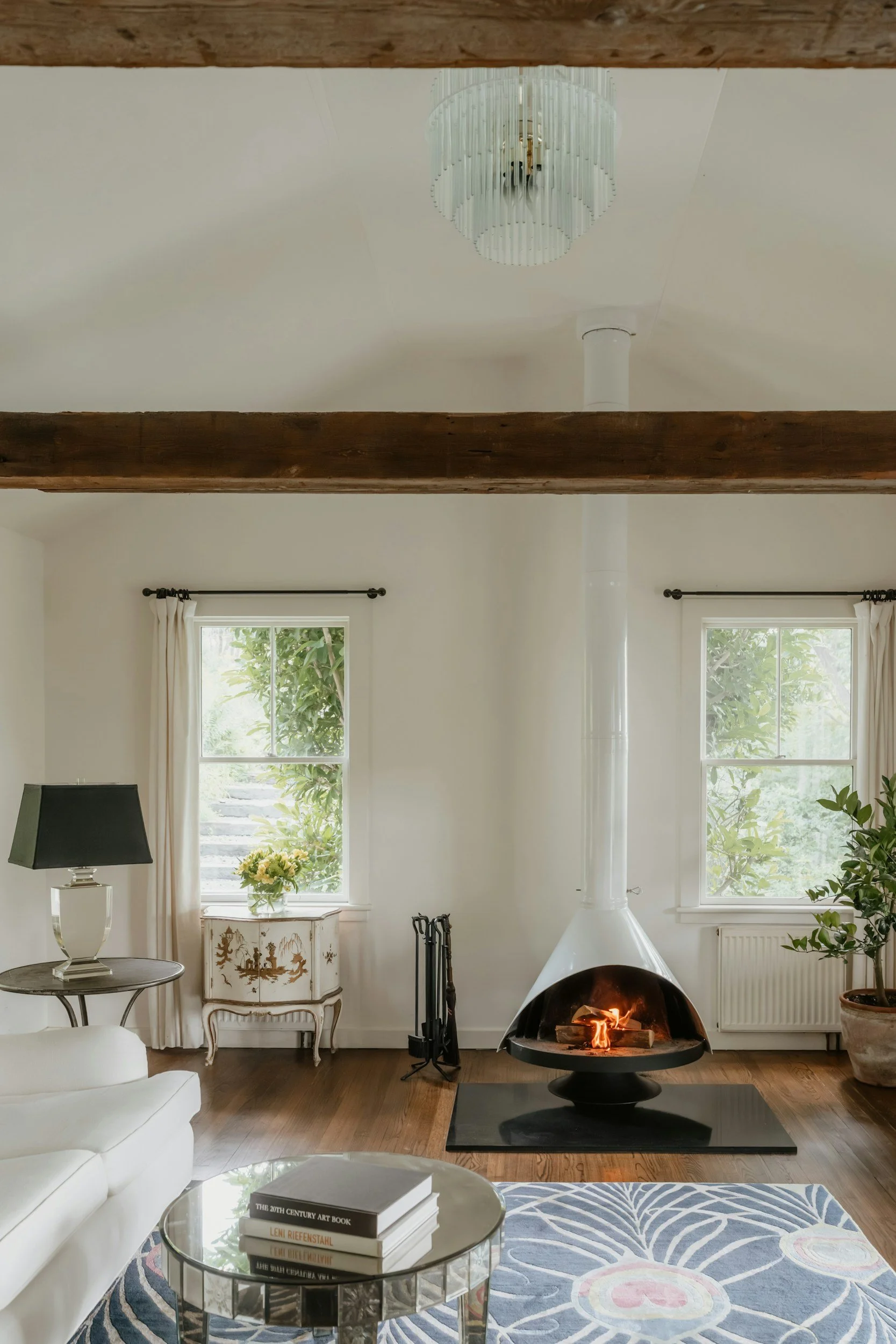 Living room with a modern fireplace, two windows with white curtains, a decorative white cabinet, a black table with books, a white sofa, a rug with a floral pattern, a black lampshade, and green plants.