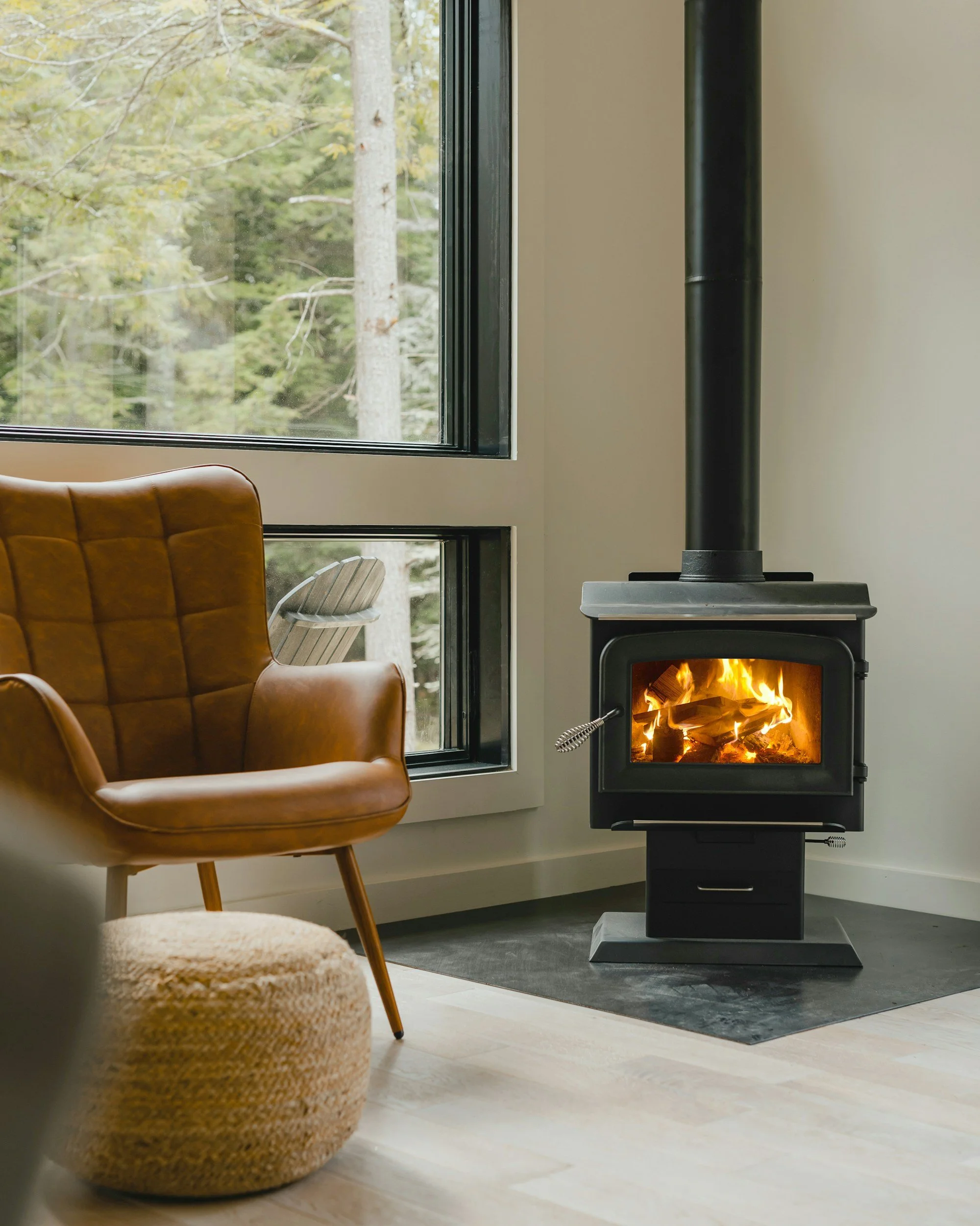 Interior of a cozy room with a leather armchair, a window overlooking a forest, and a wood-burning stove with a fire inside.