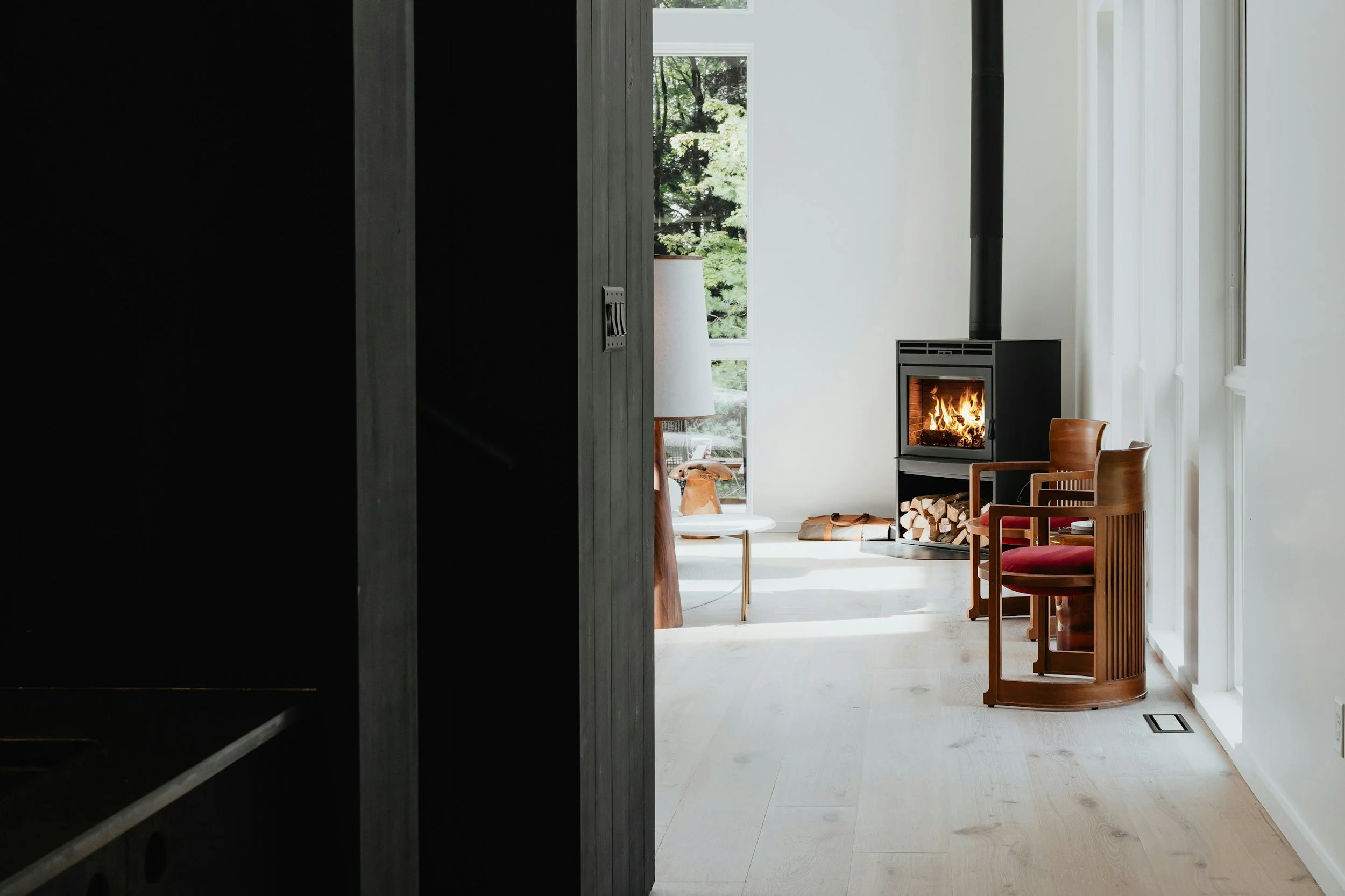 Living room with a wood-burning stove, wooden chairs with red cushions, a table, and large windows showing greenery outside.