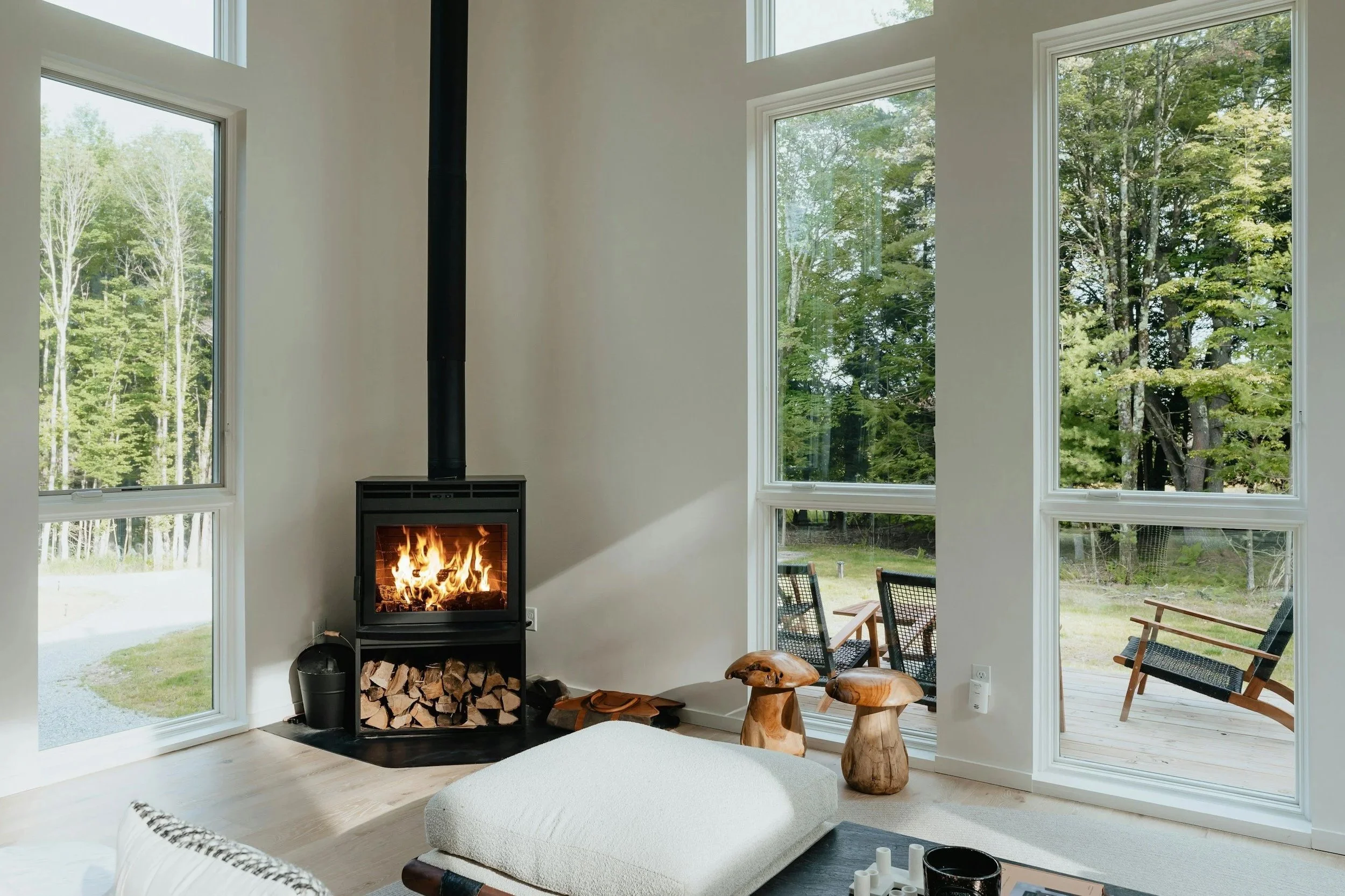 Living room with large windows showing a green forest outside, a fireplace with a fire burning, and modern furniture including a white cushioned lounge, wooden stools, and chairs on a deck.