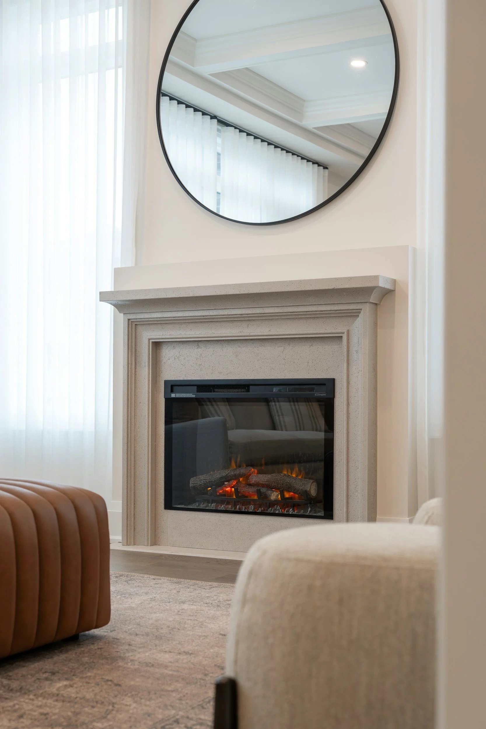 Living room with a beige stone fireplace, a round mirror above it, and a fire burning inside. Light streaming through sheer white curtains on the windows.