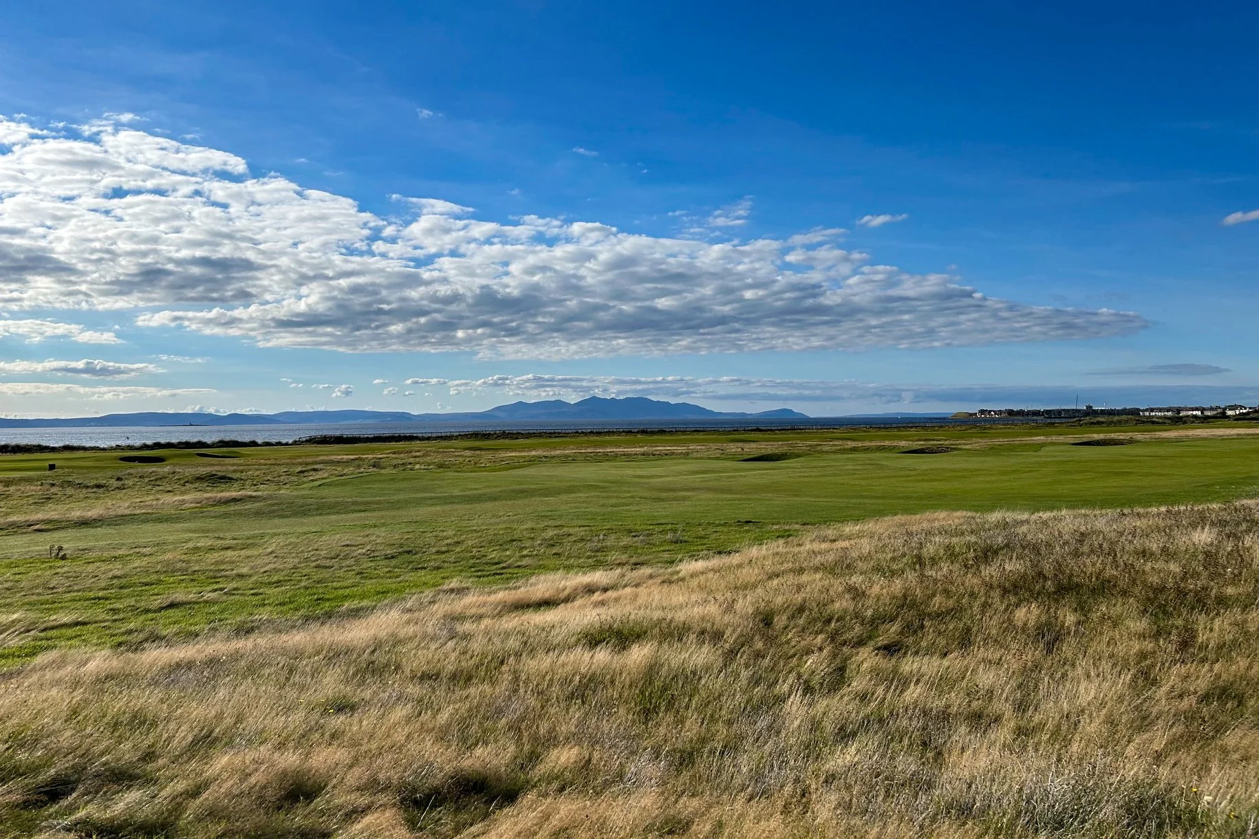 Scenic view of a golf course with lush green grass, a body of water in the distance, and mountains under a partly cloudy blue sky.