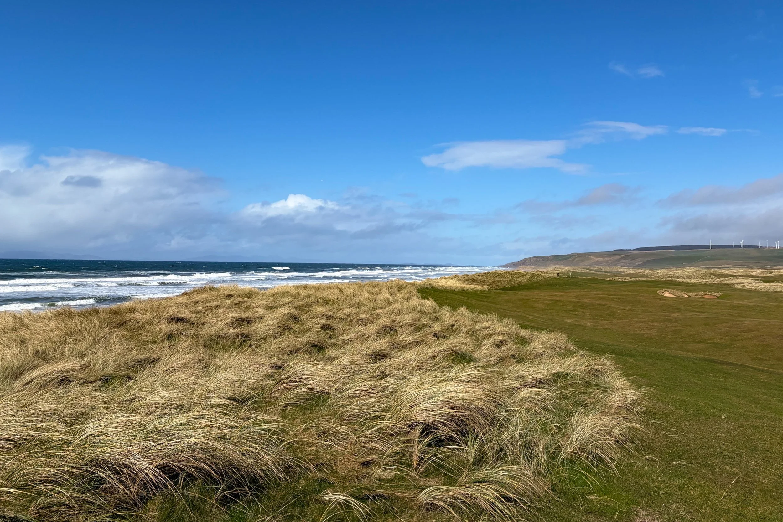 A coastal landscape with a sandy grassy foreground, ocean waves, and a distant hillside with wind turbines under a partly cloudy blue sky.