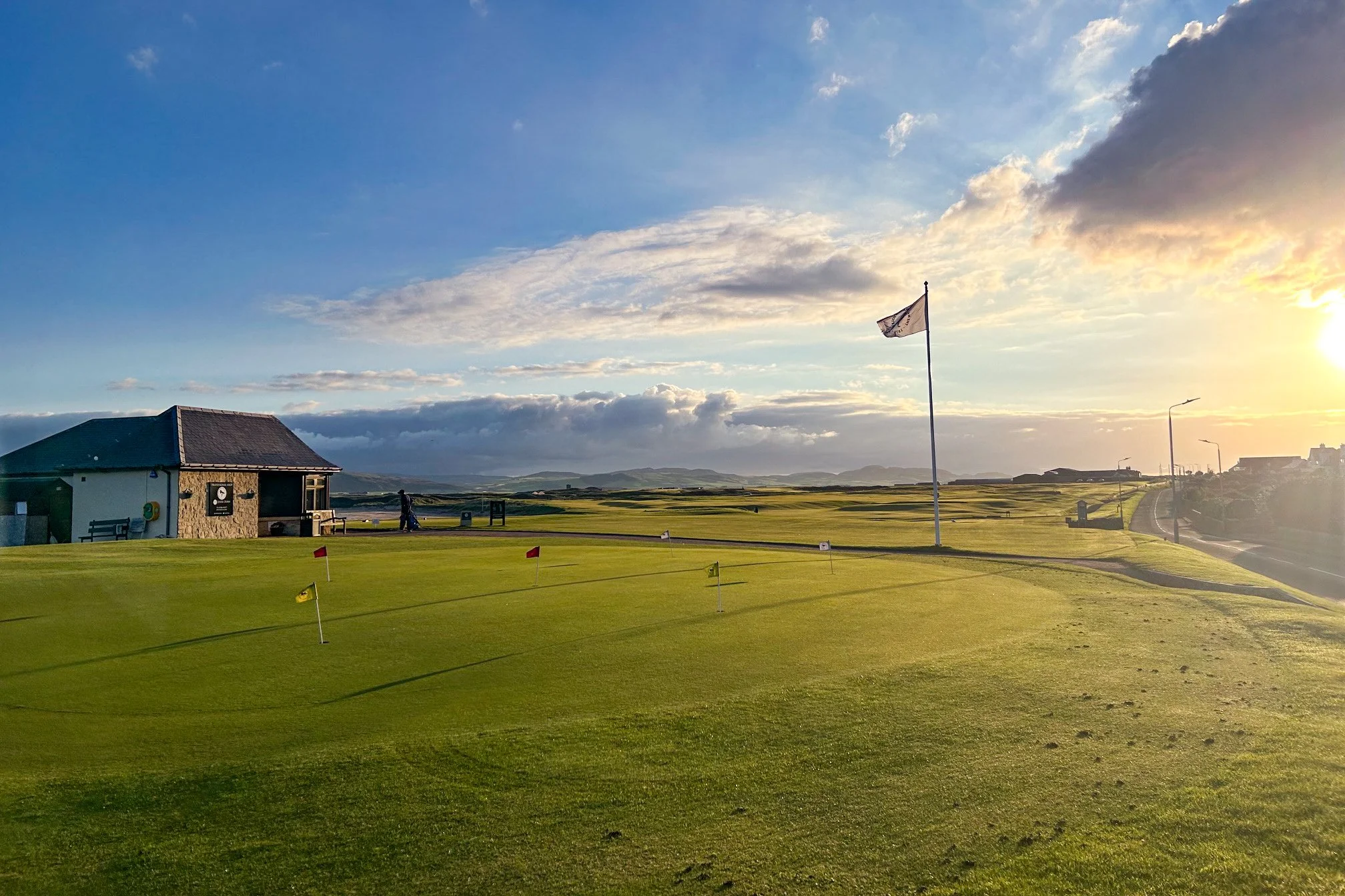 A golf course with a putting green and small flags, a stone building, a flagpole with a flag, streetlights, a pathway, and a person walking, with the sun setting and clouds in the sky.