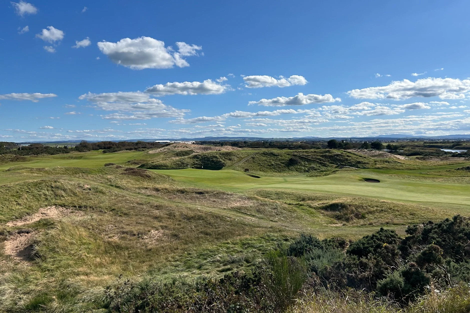 A golf course surrounded by hillocks and grass with a bright blue sky and scattered clouds overhead.