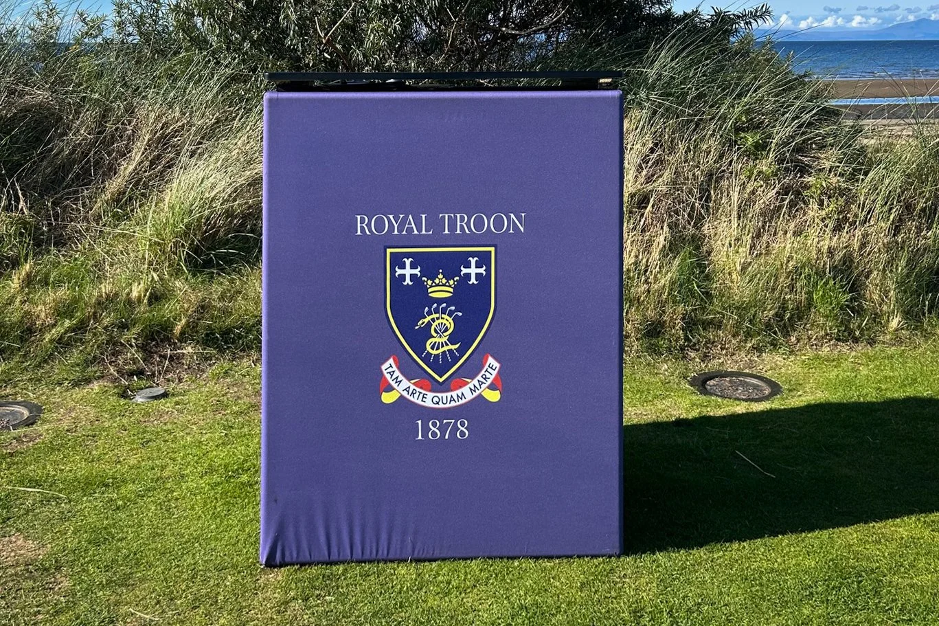 A purple sign with the Royal Troon golf club crest, including a crown, shield, and the Latin motto 'TAm ArTE qUAM MARTE,' situated outdoors on a grassy area with dunes and ocean in the background.