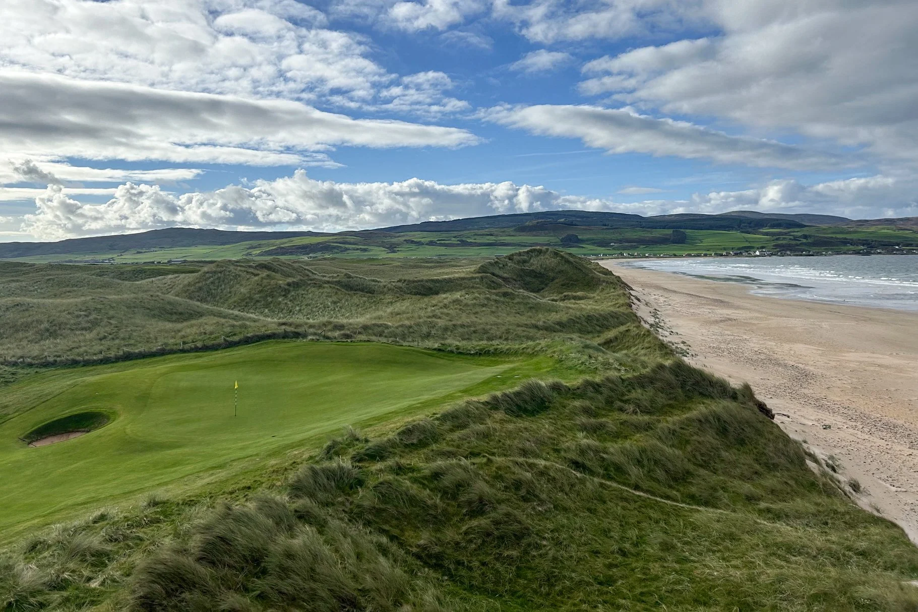 A golf course next to a sandy beach with grassy dunes and rolling green hills under a partly cloudy sky.