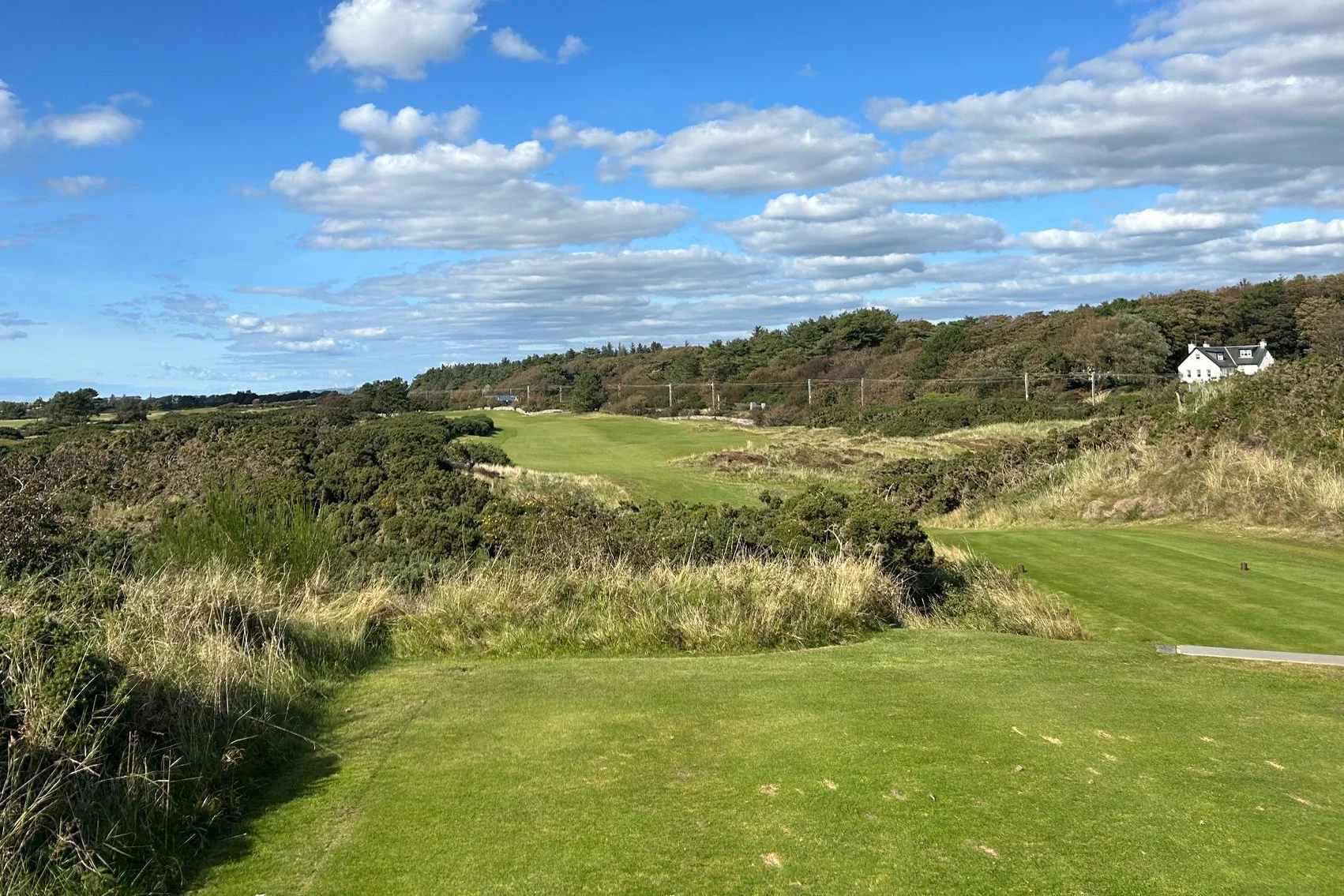 A scenic view of a golf course with green grass, surrounded by rolling hills, bushes, and trees under a partly cloudy sky.
