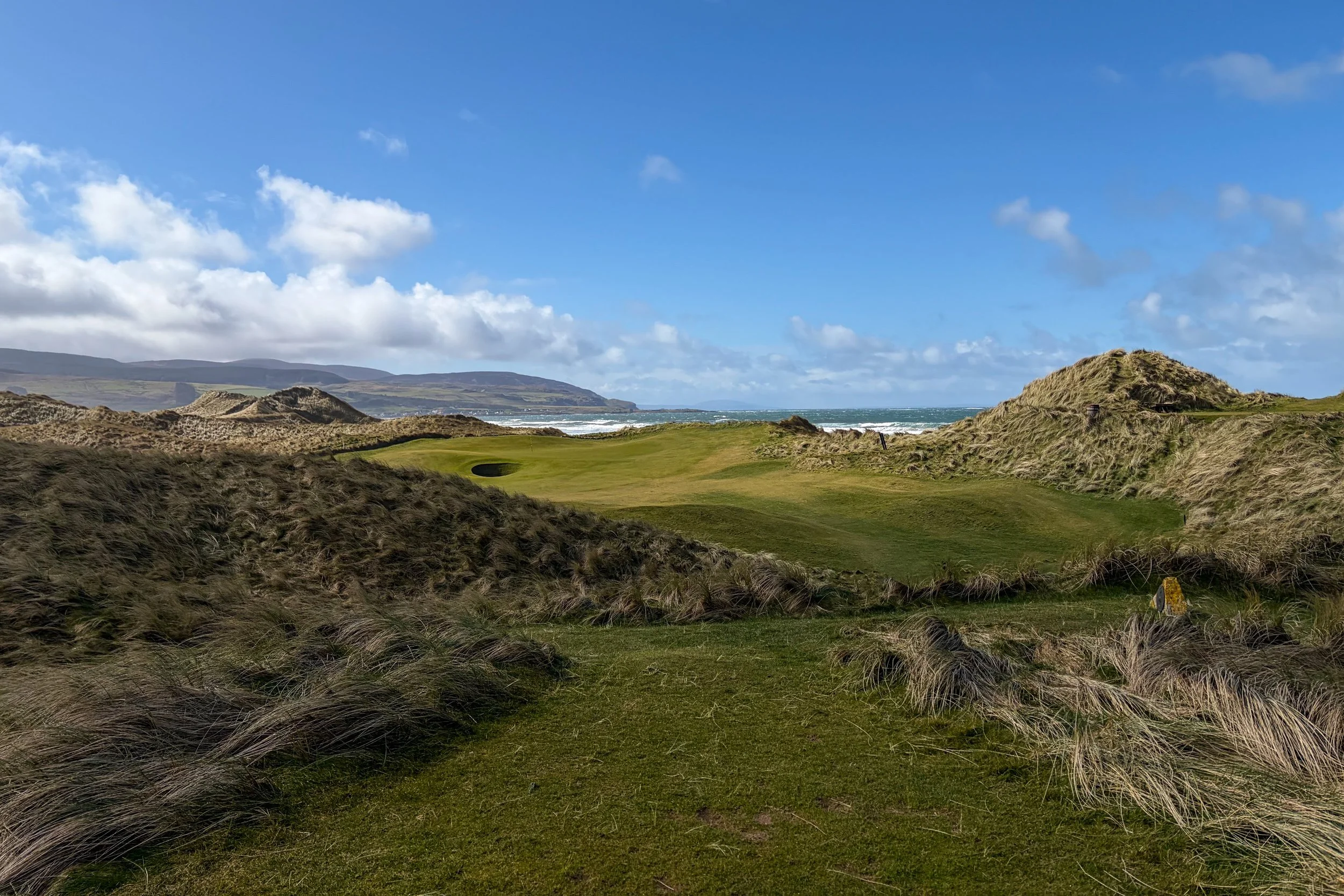 A coastal golf course with green fairways surrounded by sandy dunes and tall grass, with the ocean in the background and a partly cloudy sky above.