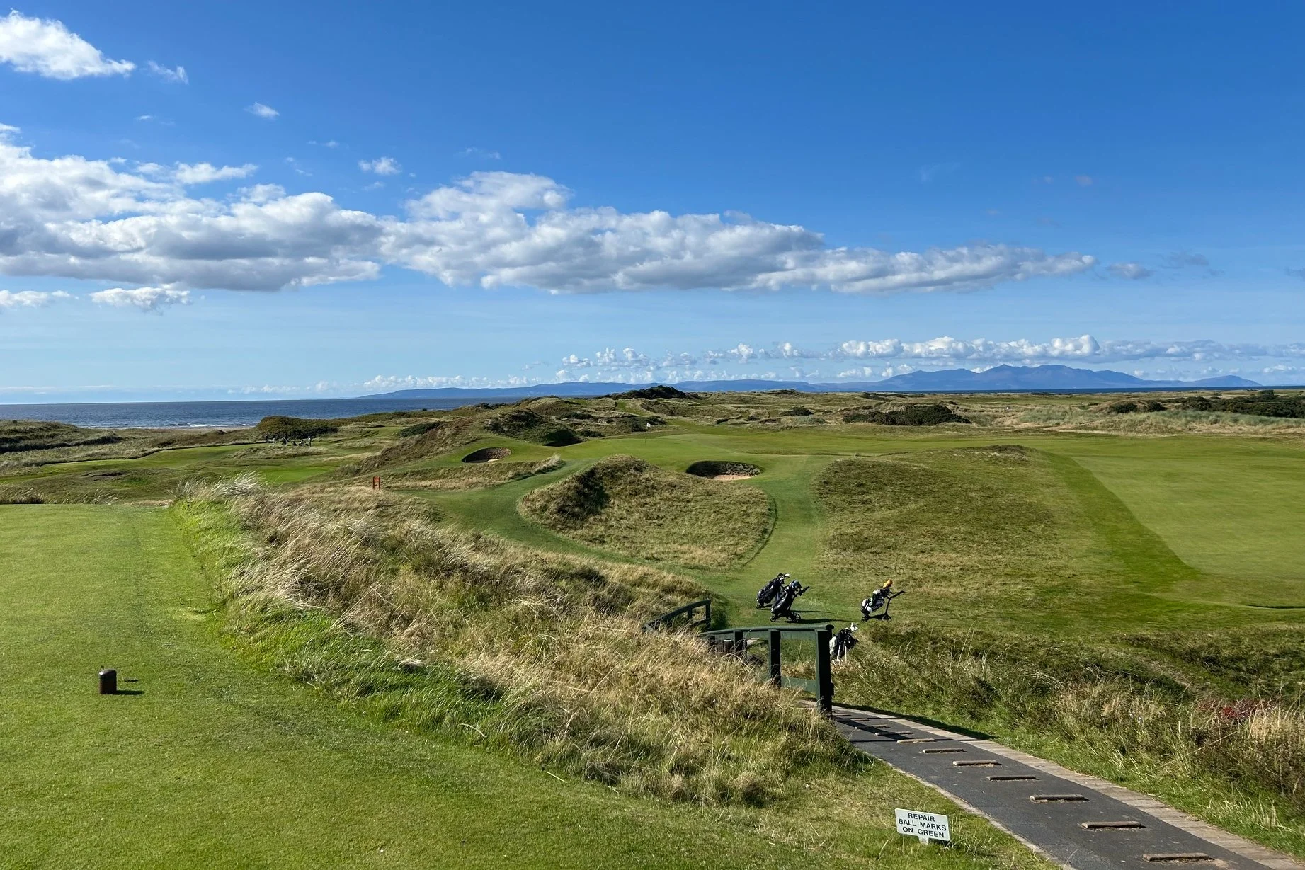A scenic golf course with lush green fairways, sand bunkers, and golf bags on cart paths, overlooking the ocean with mountains in the background under a partly cloudy sky.
