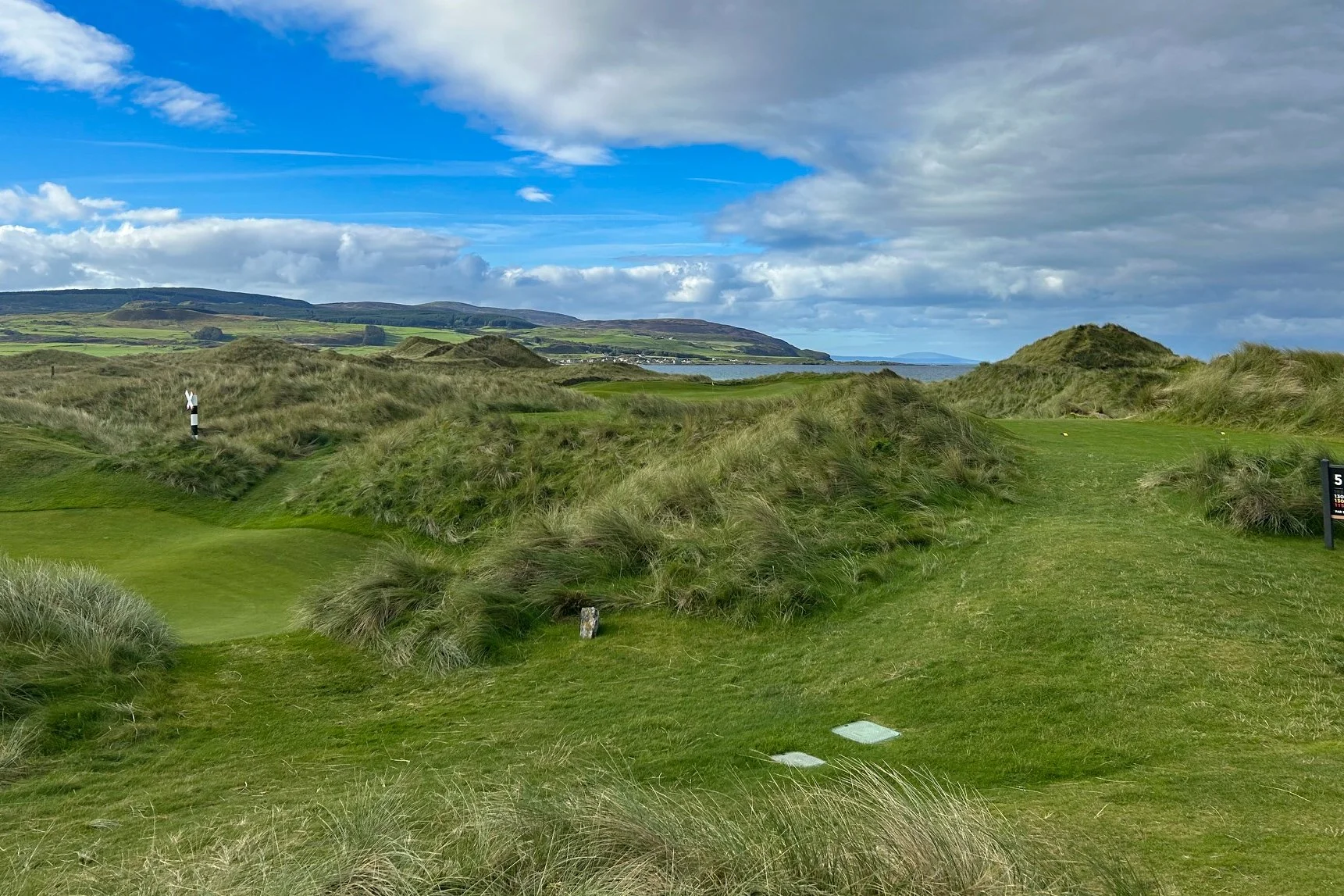 A golf course with green grass, sand dunes, and a small pond on the left. In the background, there are hills and a cloudy sky.