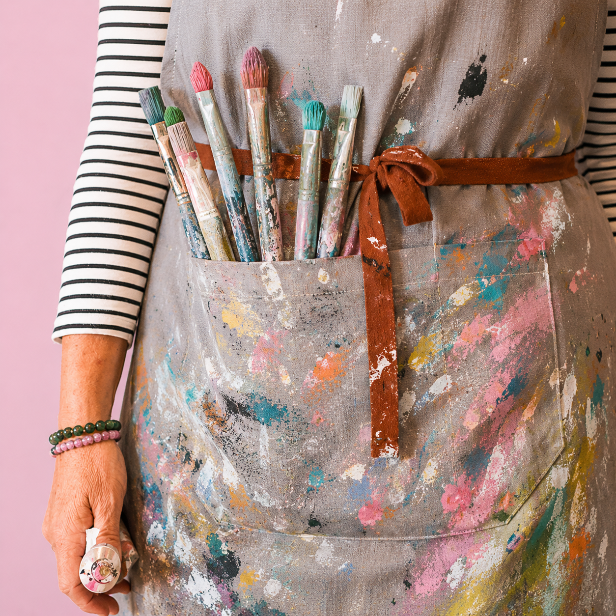 Close-up of a person wearing a paint-splattered apron with a pocket holding paintbrushes, black and white striped shirt, and pink and green bracelets.