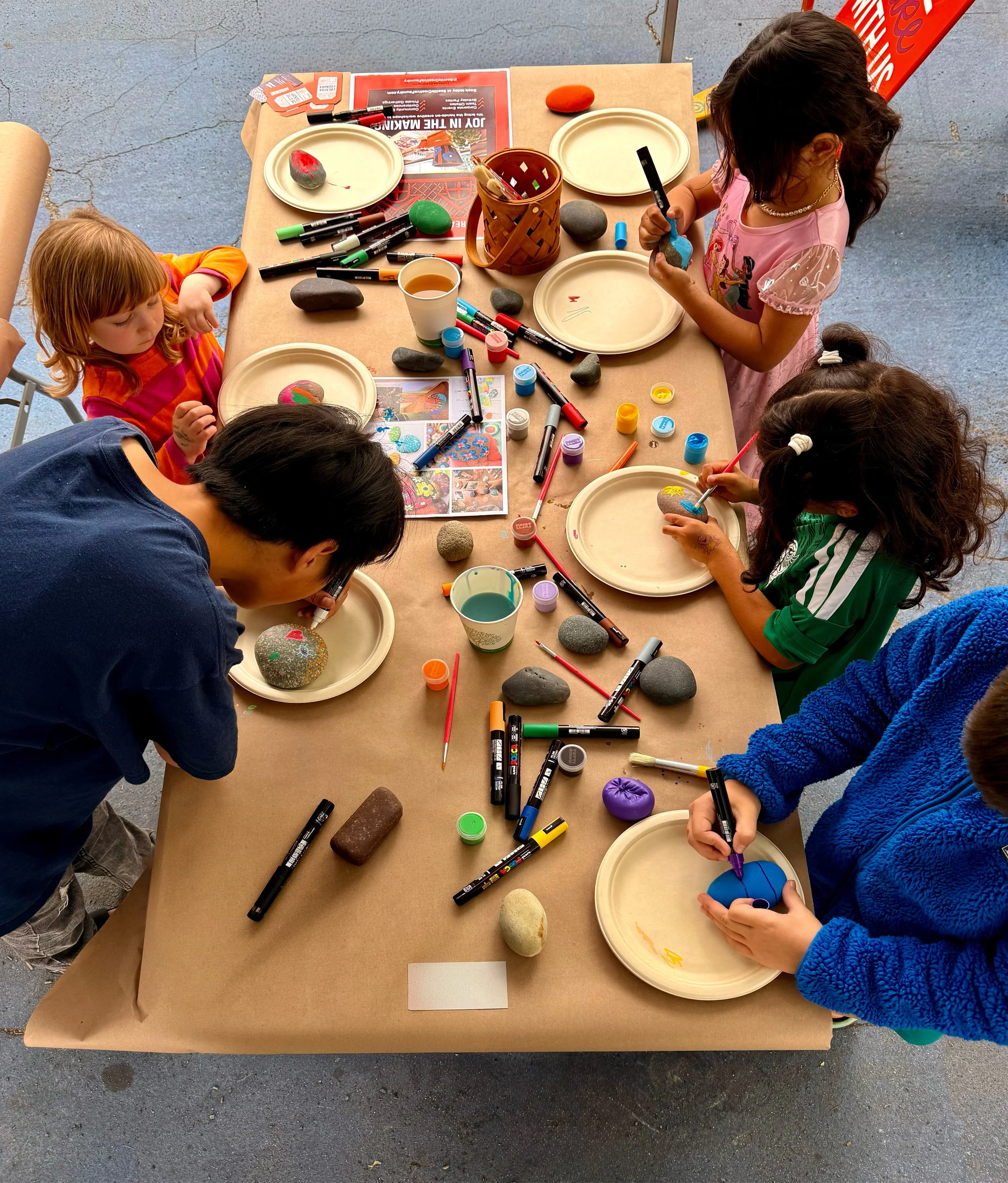 Children doing rock painting at a table with rocks, paint, and markers, outdoors.
