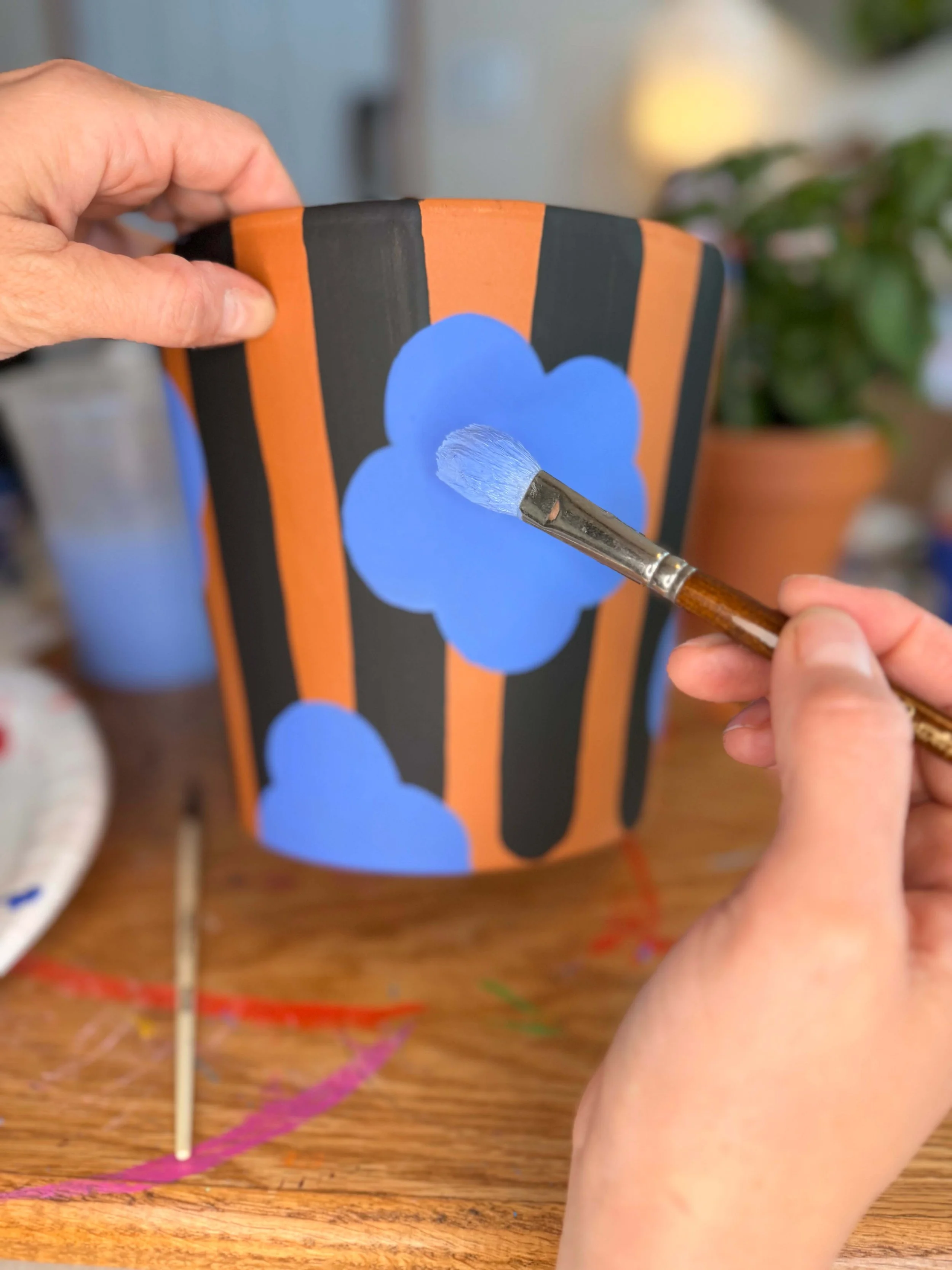 Person painting blue flowers on a painted terra cotta planter with a small brush, set on a wooden table.